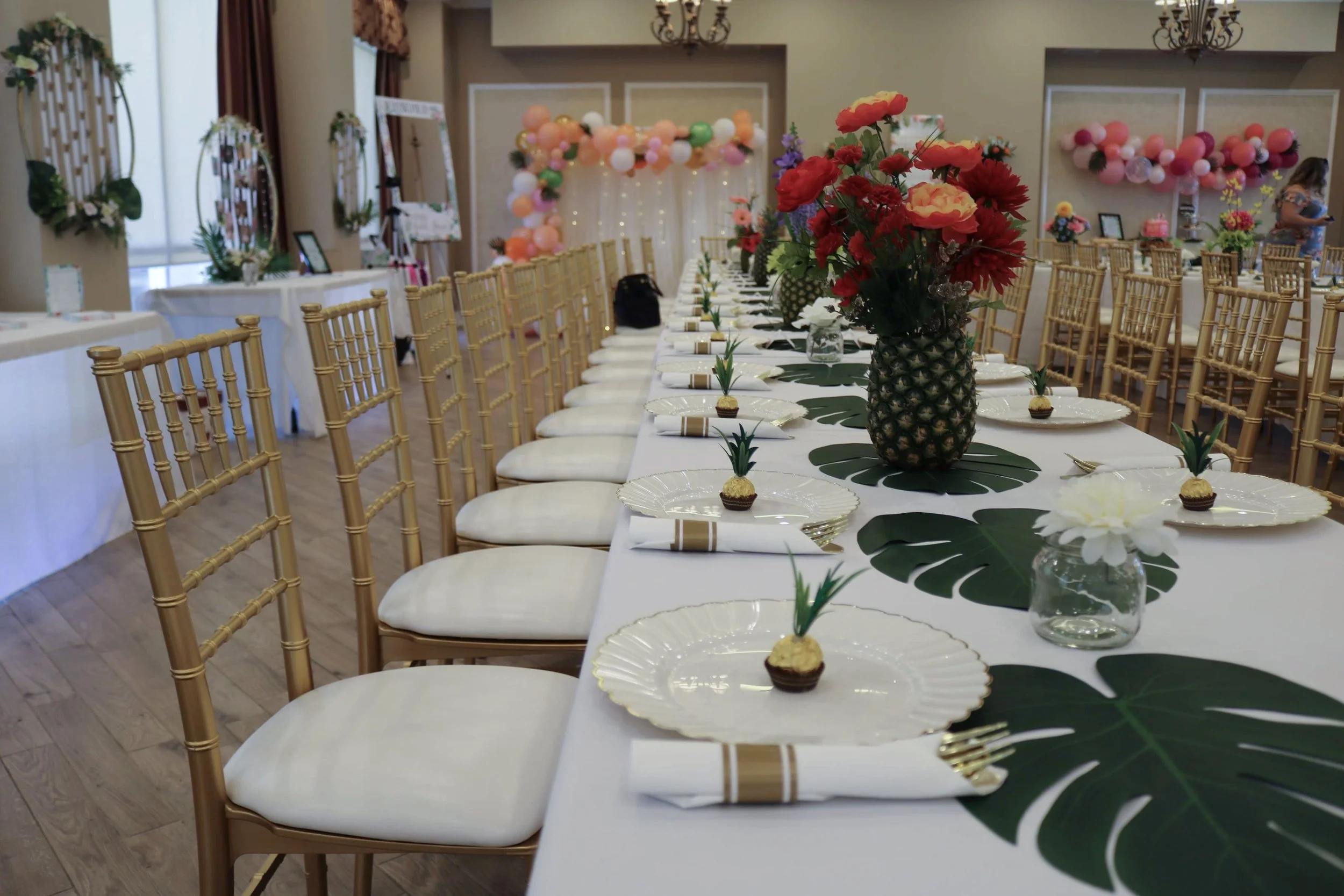 A long banquet table decorated for a celebration with a centerpiece of red and pink flowers in a pineapple-shaped vase, surrounded by small green leaves on green table runners, arranged with white plates, golden utensils, and small cupcakes with pine