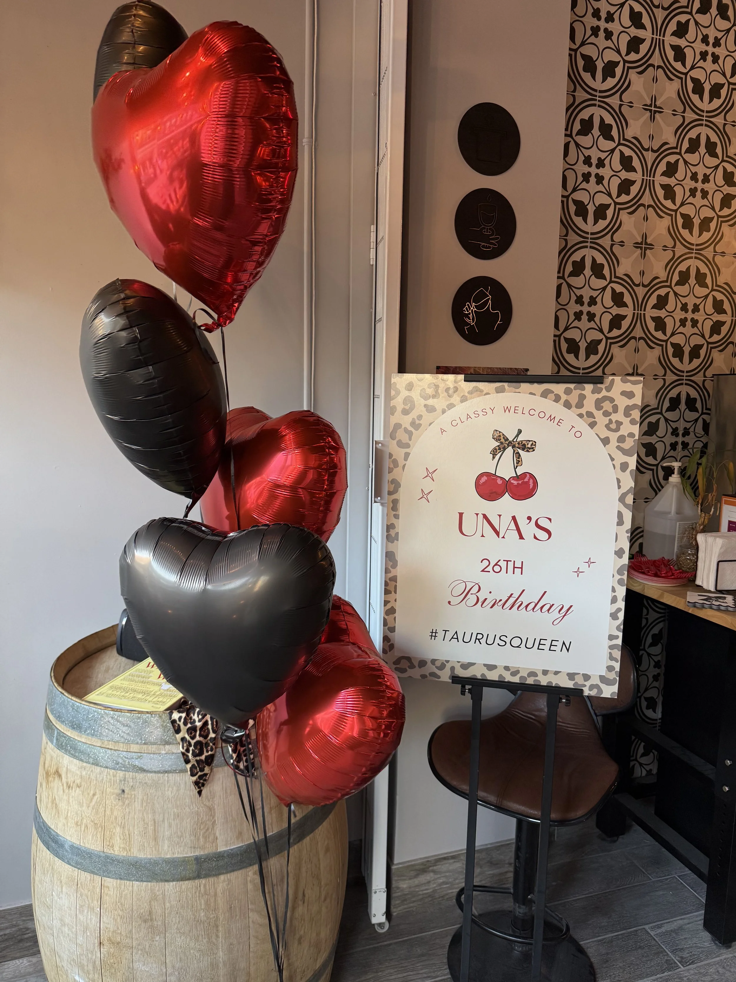 Decorative balloons in red, black, and silver are displayed beside a birthday sign for UNA'S 26th birthday, with a hashtag #TAURUSQUEEN. The setting appears to be a celebration area with a patterned wall and a table.