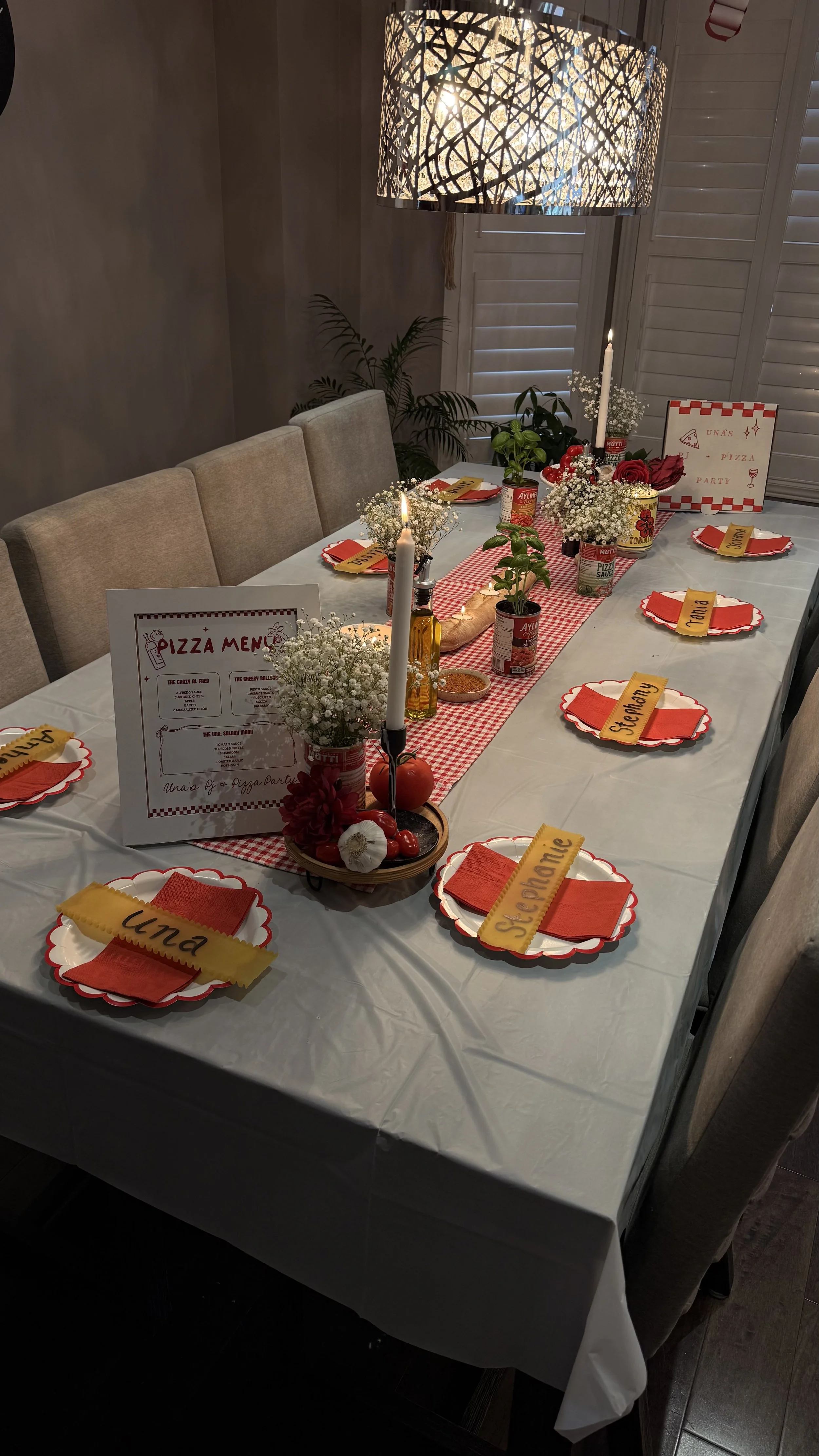 A dining table decorated for a pizza party with plates labeled with names, a pizza menu, candles, flowers, and canned tomatoes. The table has a red and white checkered table runner, and there are window shutters in the background.