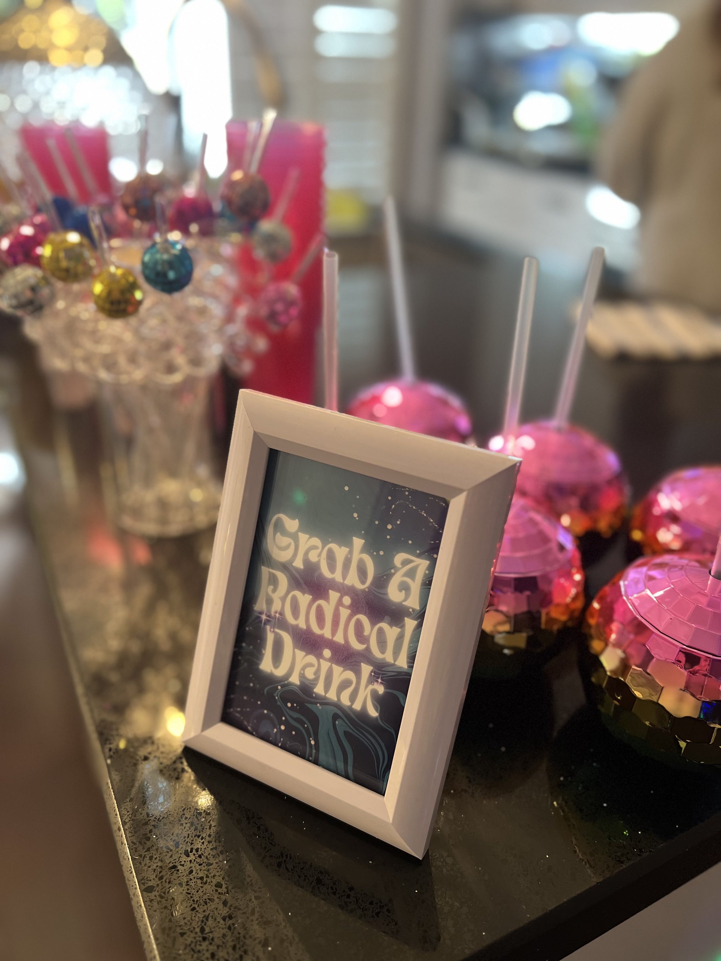 A small framed sign on a countertop reads, 'Grab A Radicad Drink.' Behind it are pink and colorful decorative items, including lollipops and shiny, multicolored spheres.