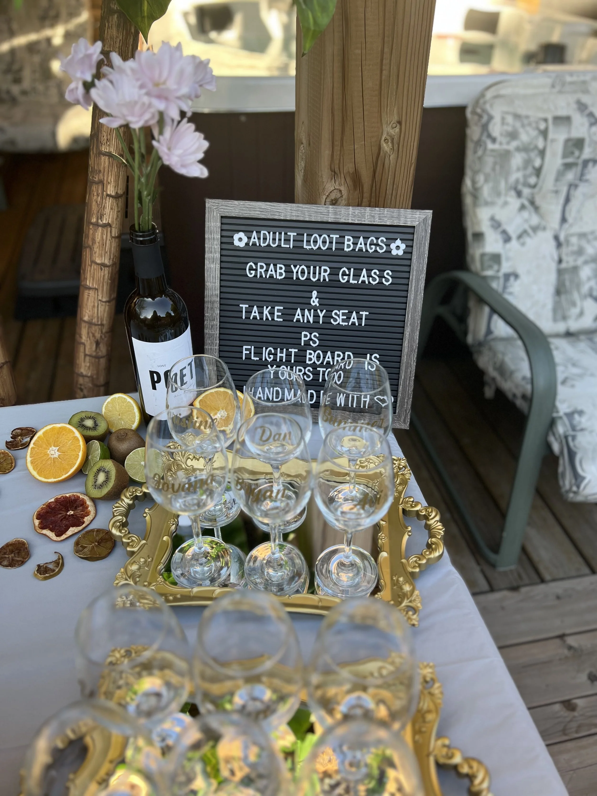 A table with a tray of sliced citrus fruits and kiwi, empty glasses, and a bottle of alcohol. A sign with rules for a party or event is behind the table. The setting appears to be outdoors, possibly on a balcony or deck.