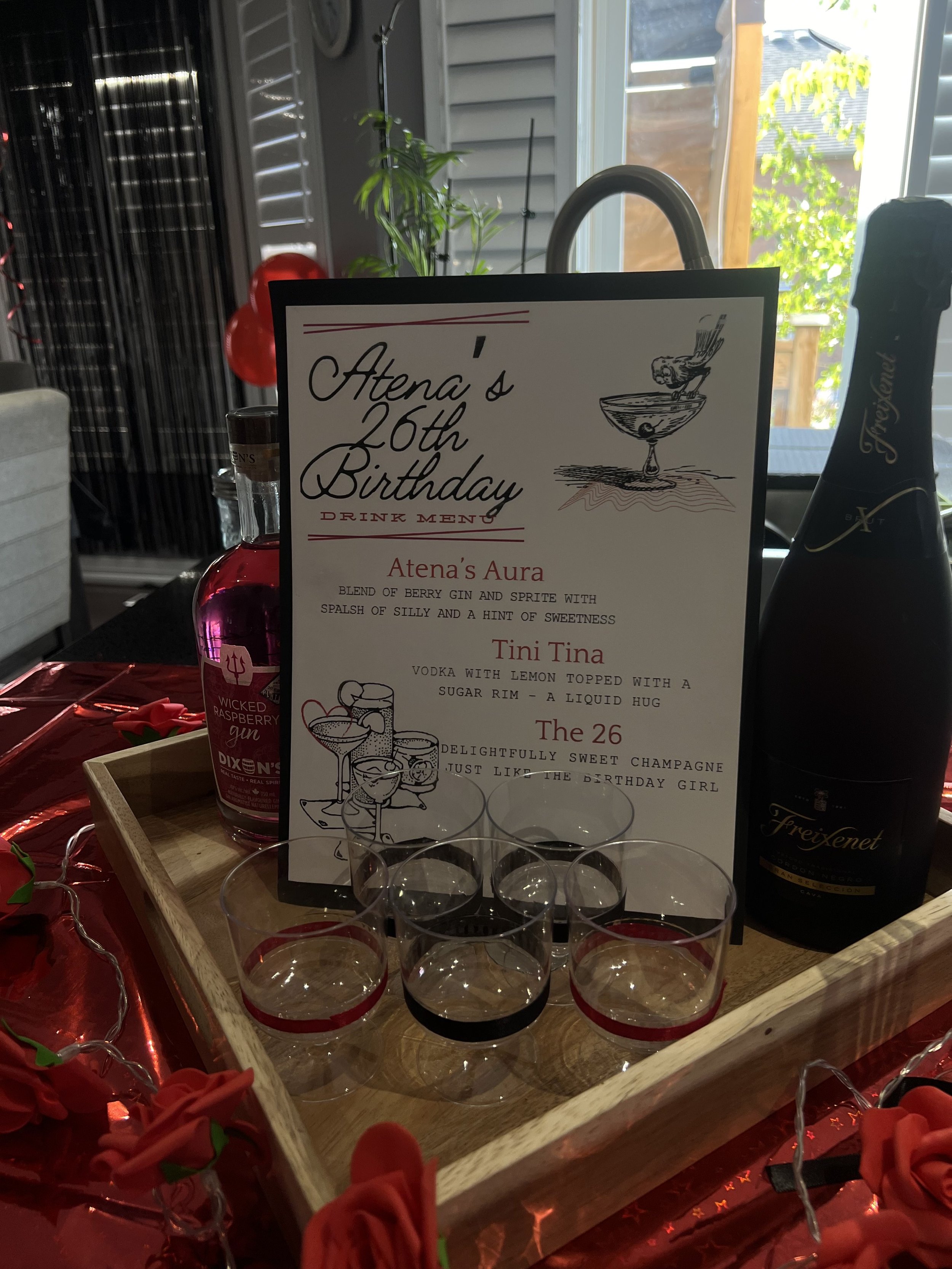 A display of adult shot glasses with a decorative sign for a 26th birthday celebration. The sign features hand-drawn illustrations of a cocktail glass and a bird, with descriptions of themed drinks. The background includes a bottle of flavored gin an
