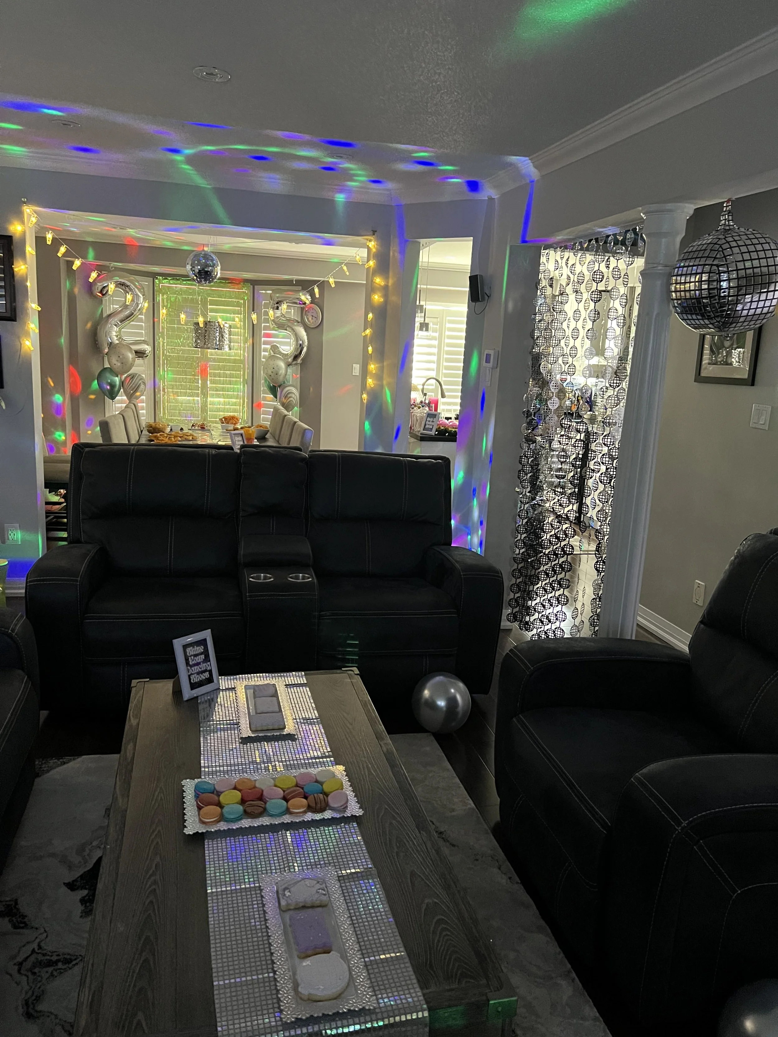 A living room decorated for a celebration with balloons, a disco ball, and colorful lights. There is a dining table in the background and a black couch in the foreground. On the table, there are macarons and a fan of baked goods. The room has string 
