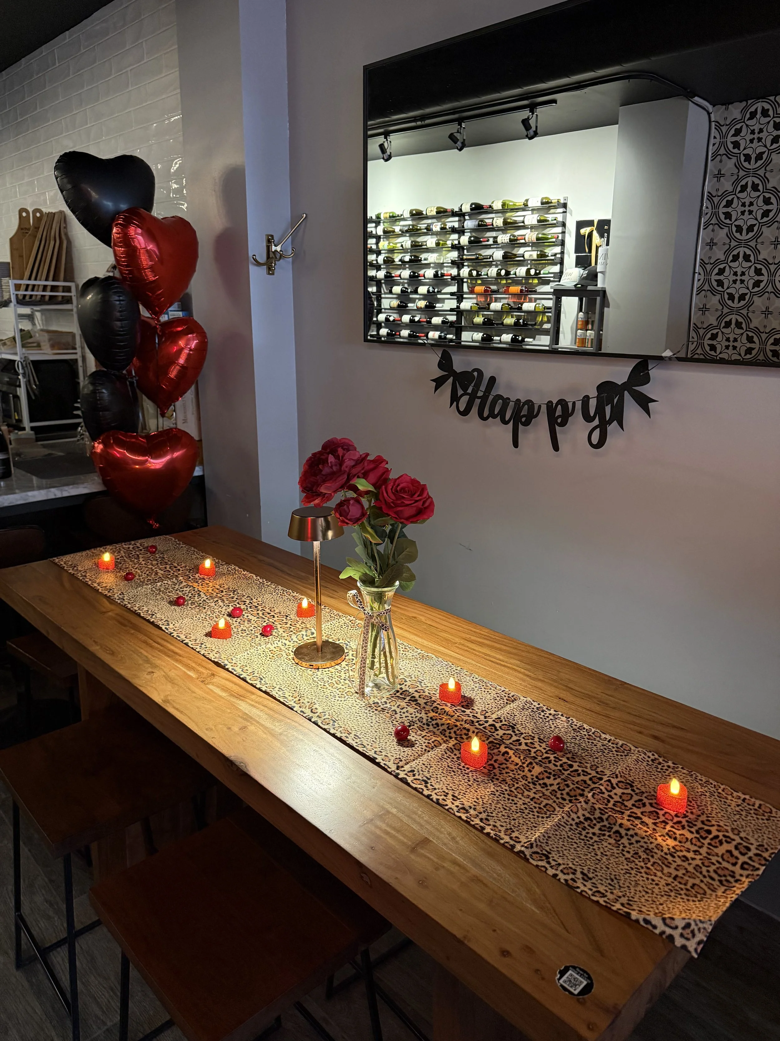 A dining table decorated with red and black heart-shaped balloons on the left, a vase with pink roses, small candles, and red decorative accents; a leopard print table runner and a 'Happy' banner on the wall. A mirror reflects a wine rack and a cabin