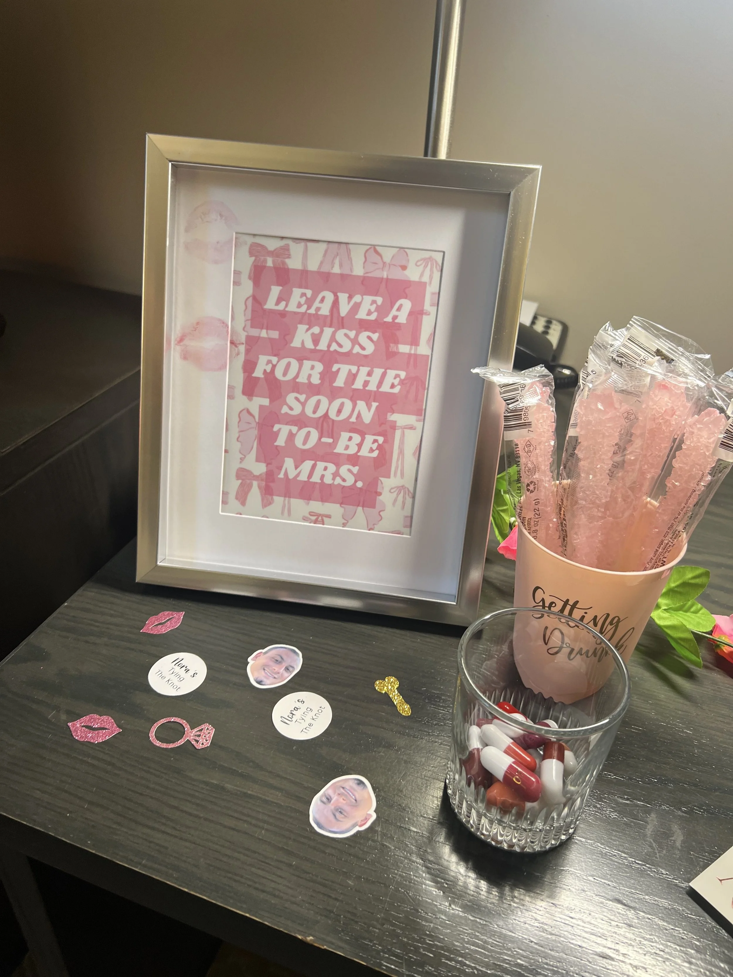 A framed sign with pink and white text saying 'Leave a kiss for the soon-to-be Mrs.' A pink container with pink rock candies and a clear cup with capsules and pills are on a dark wooden table, along with paper cutouts of lips and photos.