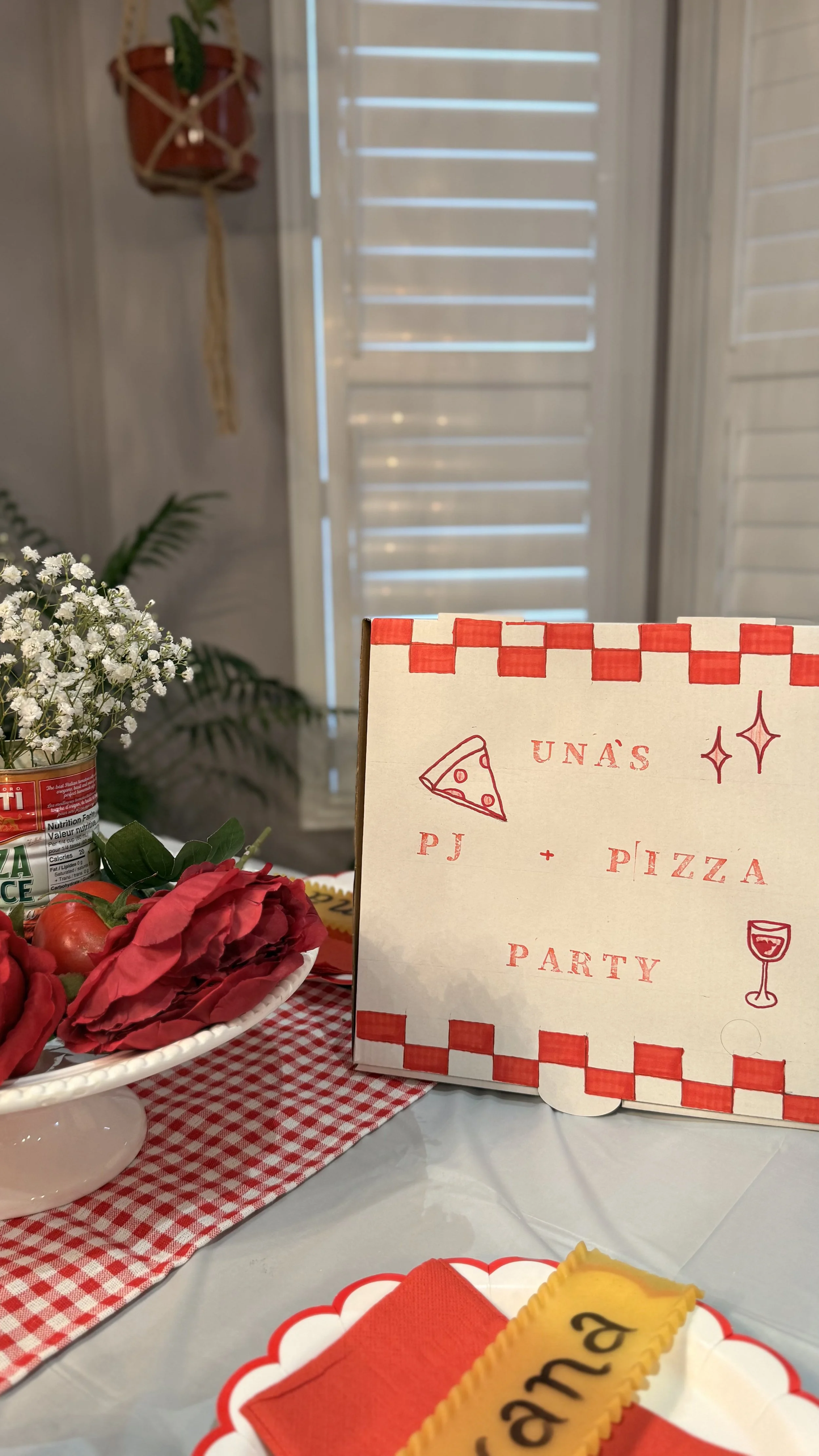 Decorative party table setup with a red and white checkered tablecloth, flowers, a pizza themed sign, and a decorated plate with a yellow and red napkin and a cookie with the word "BAM!" written on it.