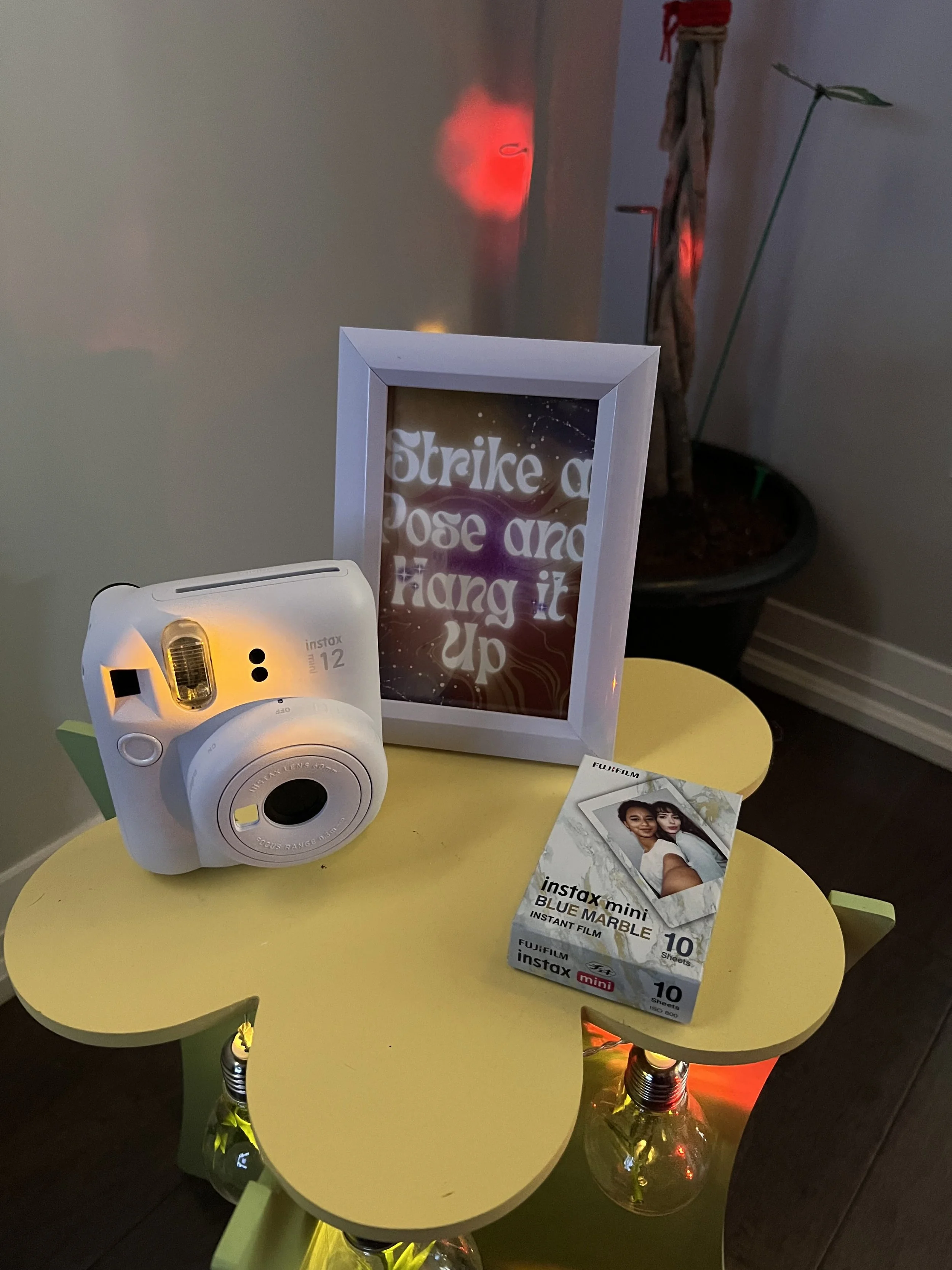 A yellow round table with a white instant camera, a box of Fujifilm Instax Mini Blue Marble instant film, a framed quote, and a small photograph of two women. A clear glass bottle with yellow and red lights inside is beneath the table. In the backgro