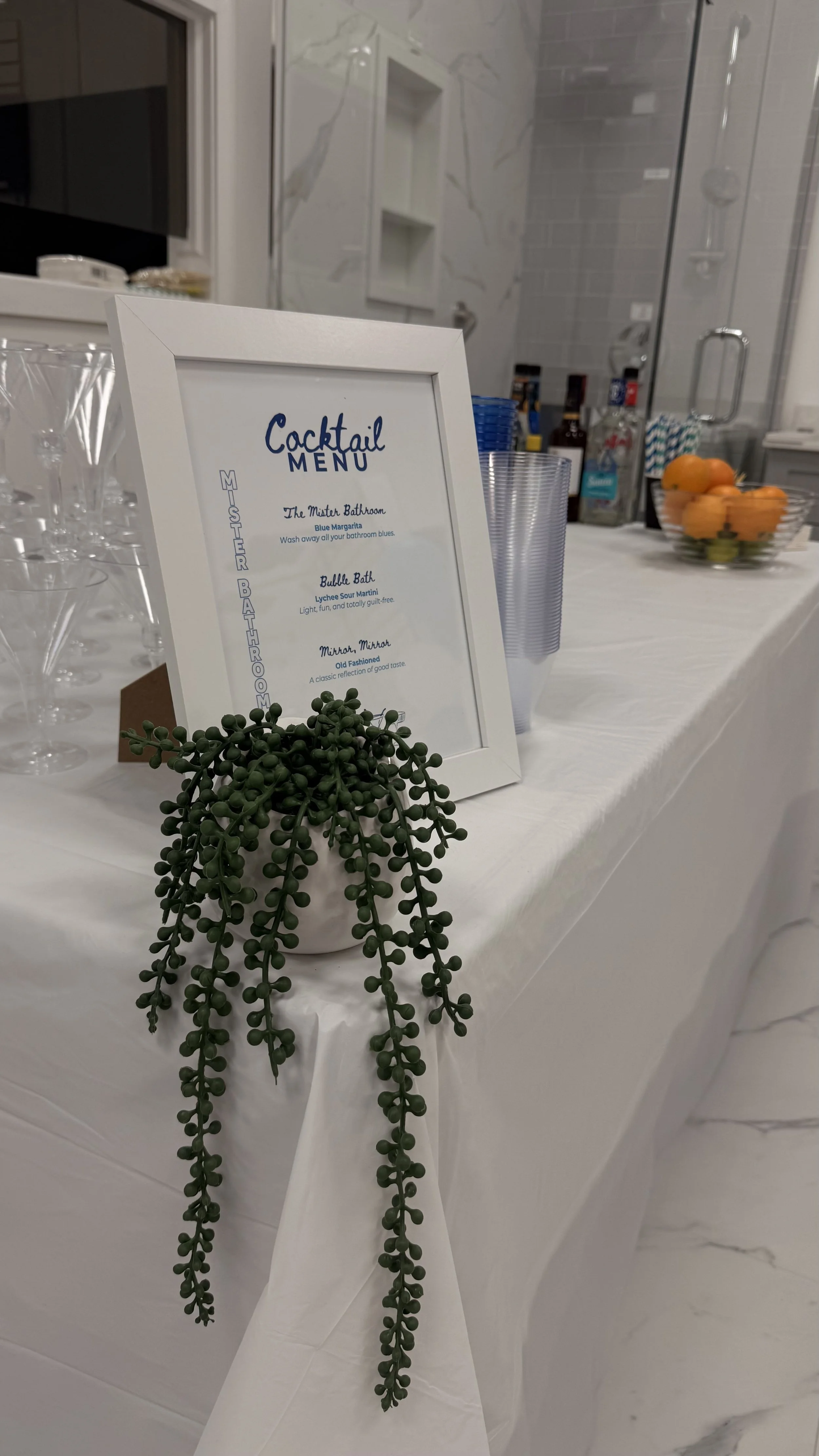 A cocktail menu on a white table with a small green plant in a white pot in front, and a clear plastic cup behind. The menu lists three drinks: Blue Margarita, Lychee Sour Martini, and Old Fashioned. In the background, there is a bowl of oranges and 