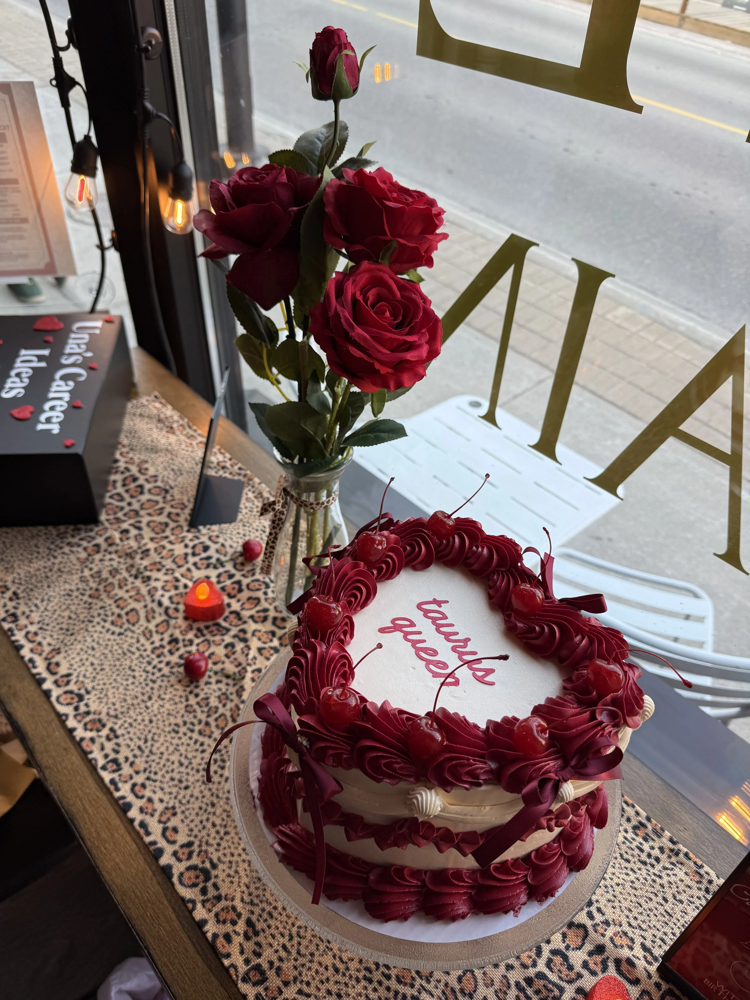 A bouquet of red roses and a cake with red frosting and cherries, with a 'Thank You' message, on a leopard-print table in a bakery window.
