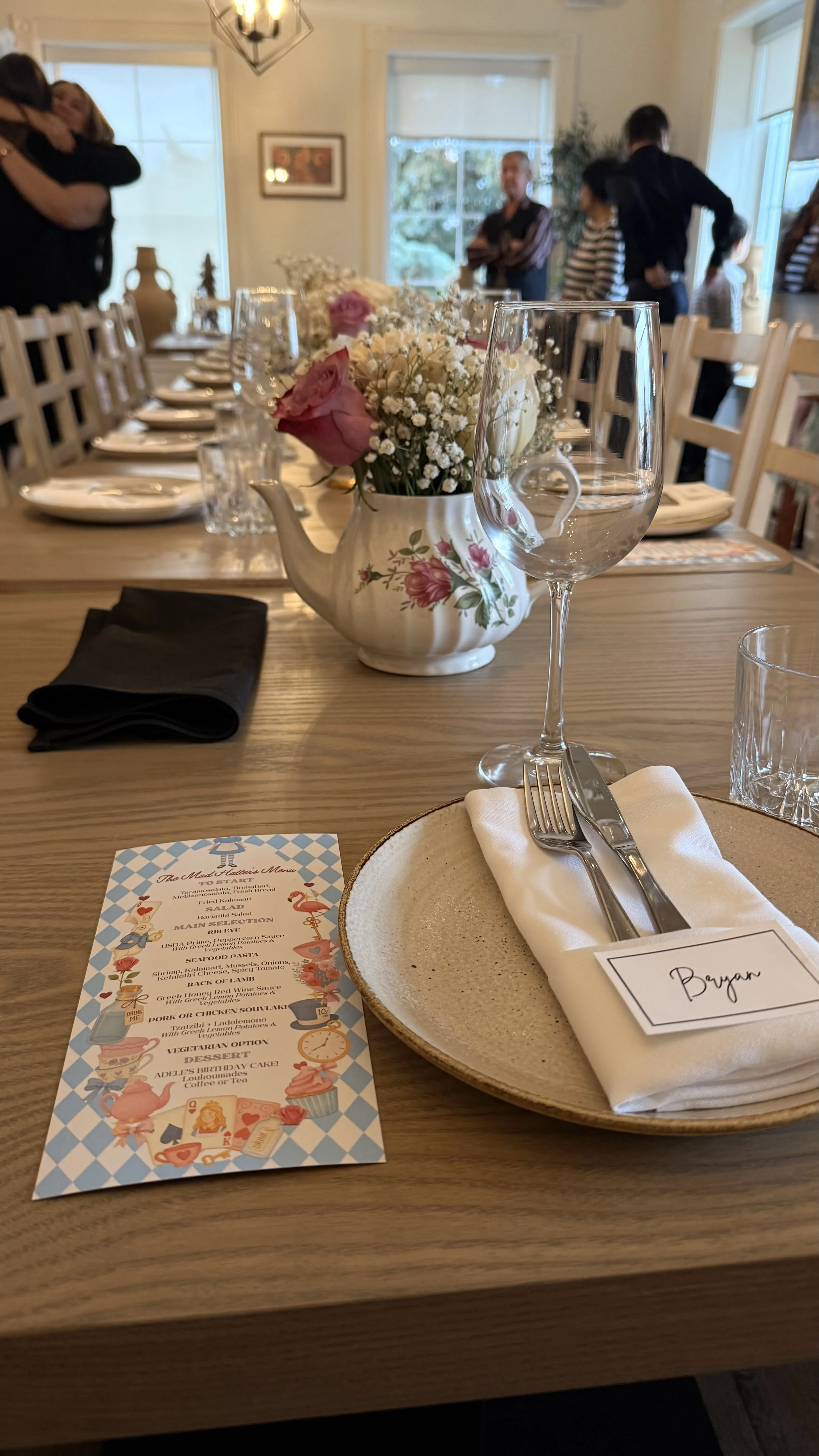 A dining table set with a floral teapot holding pink and white flowers, wine glasses, a plate with a napkin, and silverware. In the background, people are standing and hugging near windows and a picture frame.