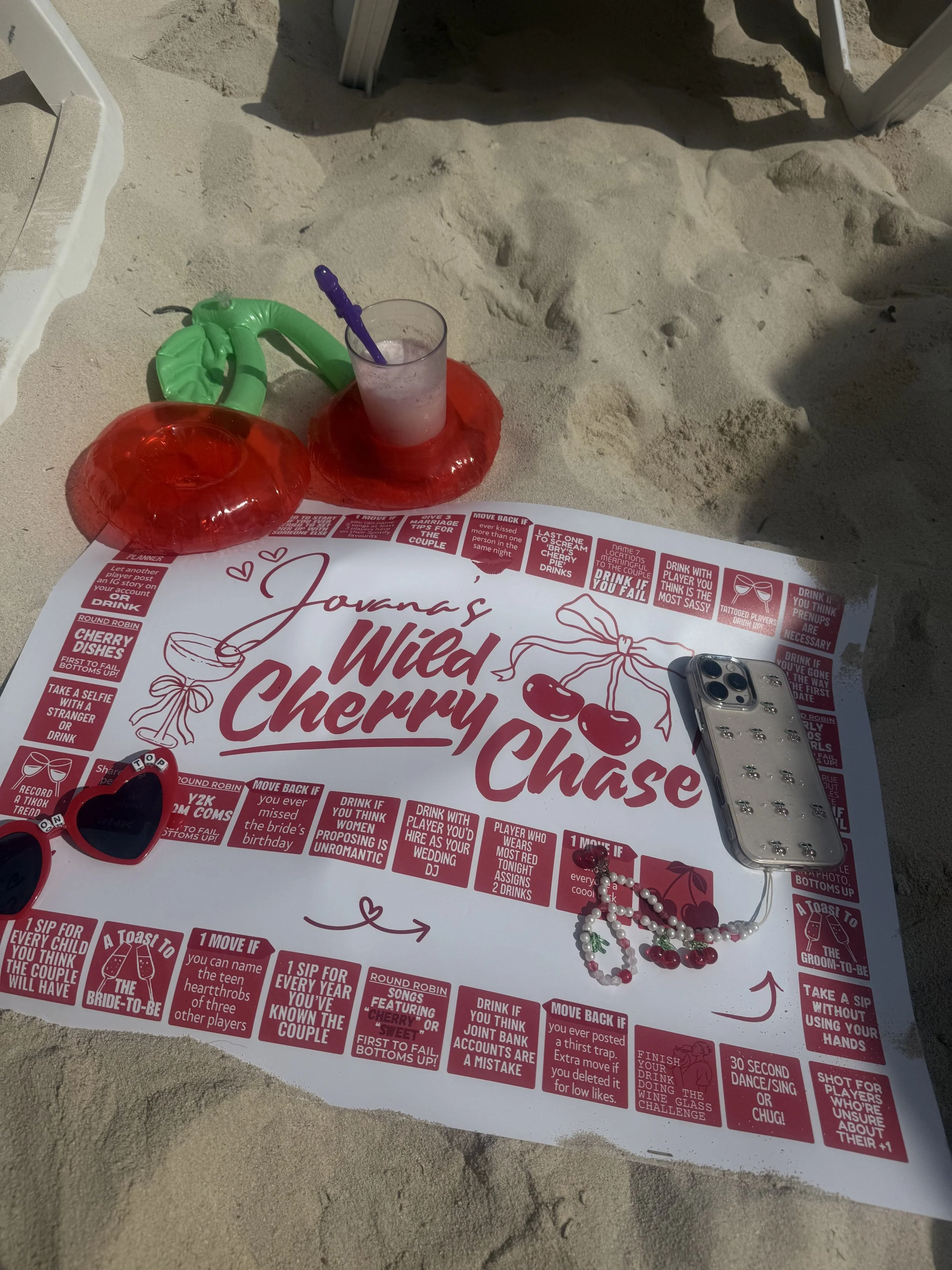 Close-up of a beach game board titled 'Johanna's Wicked Cherry Chaser,' with various playing pieces, including heart-shaped glasses, a beaded necklace, and a smartphone, situated on sandy ground.