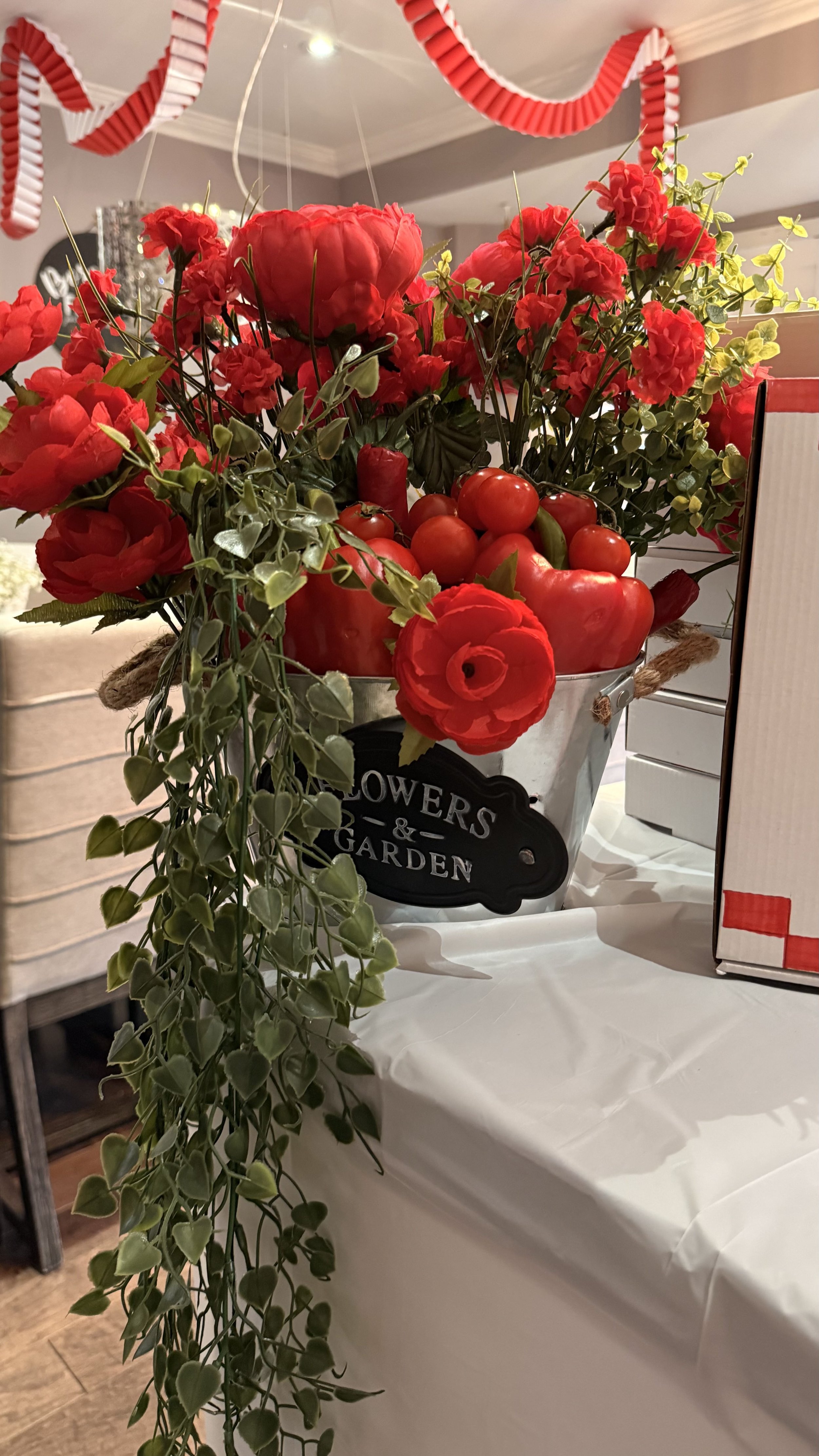 Red flowers and tomatoes arranged in a decorative container labeled "Flowers & Garden", displayed on a table with white tablecloth, in an indoor setting with red and white decorations hanging from the ceiling.