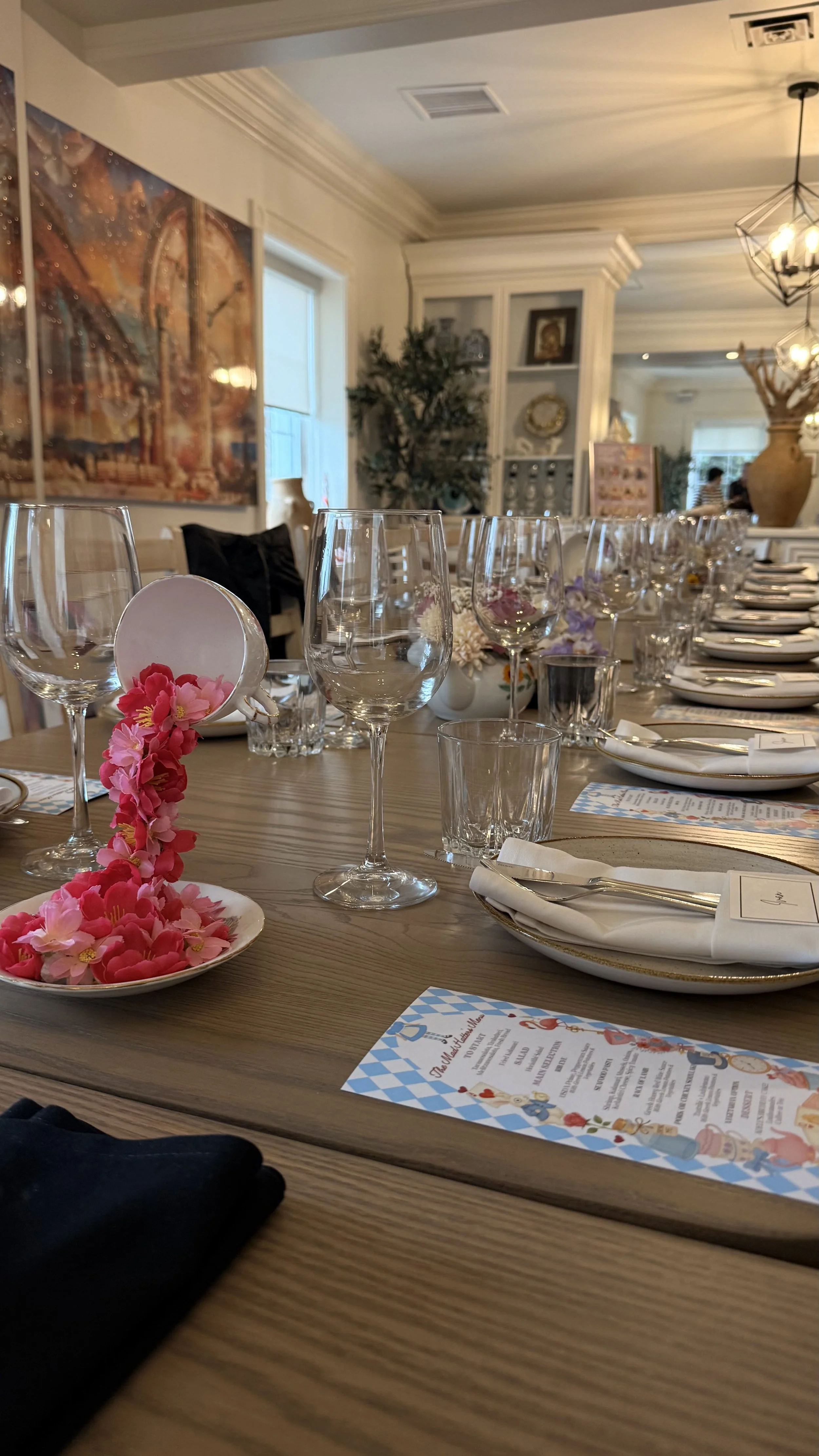 Table set for a gathering with floral centerpieces, wine glasses, plates, cutlery, and menus in an elegant room with artwork and decor.