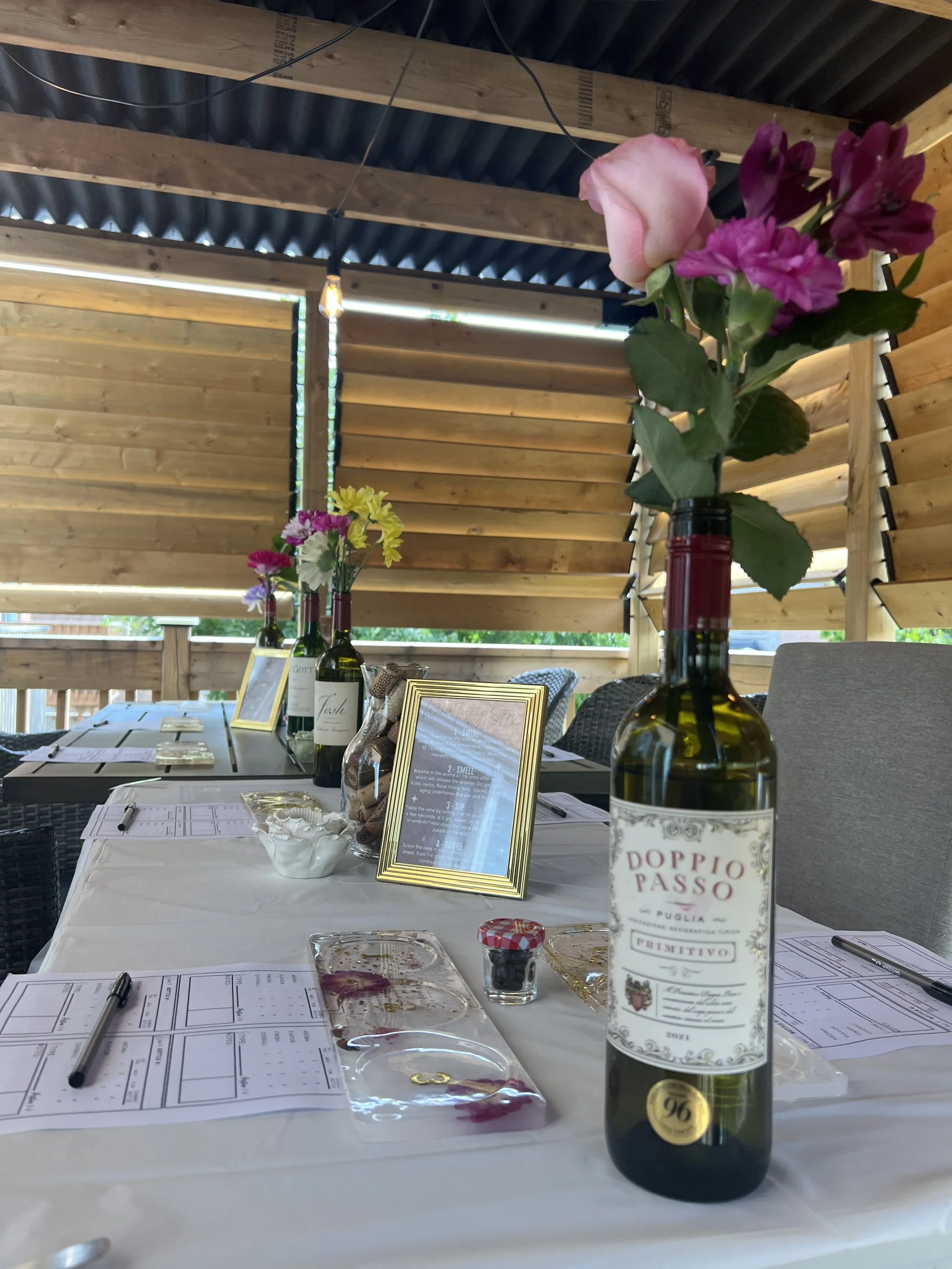 A table set for a wine tasting event with open bottles of wine, printed tasting sheets, pens, and small bowls, decorated with flowers in bottles, inside a wooden structure with horizontal slats and a dark ceiling.