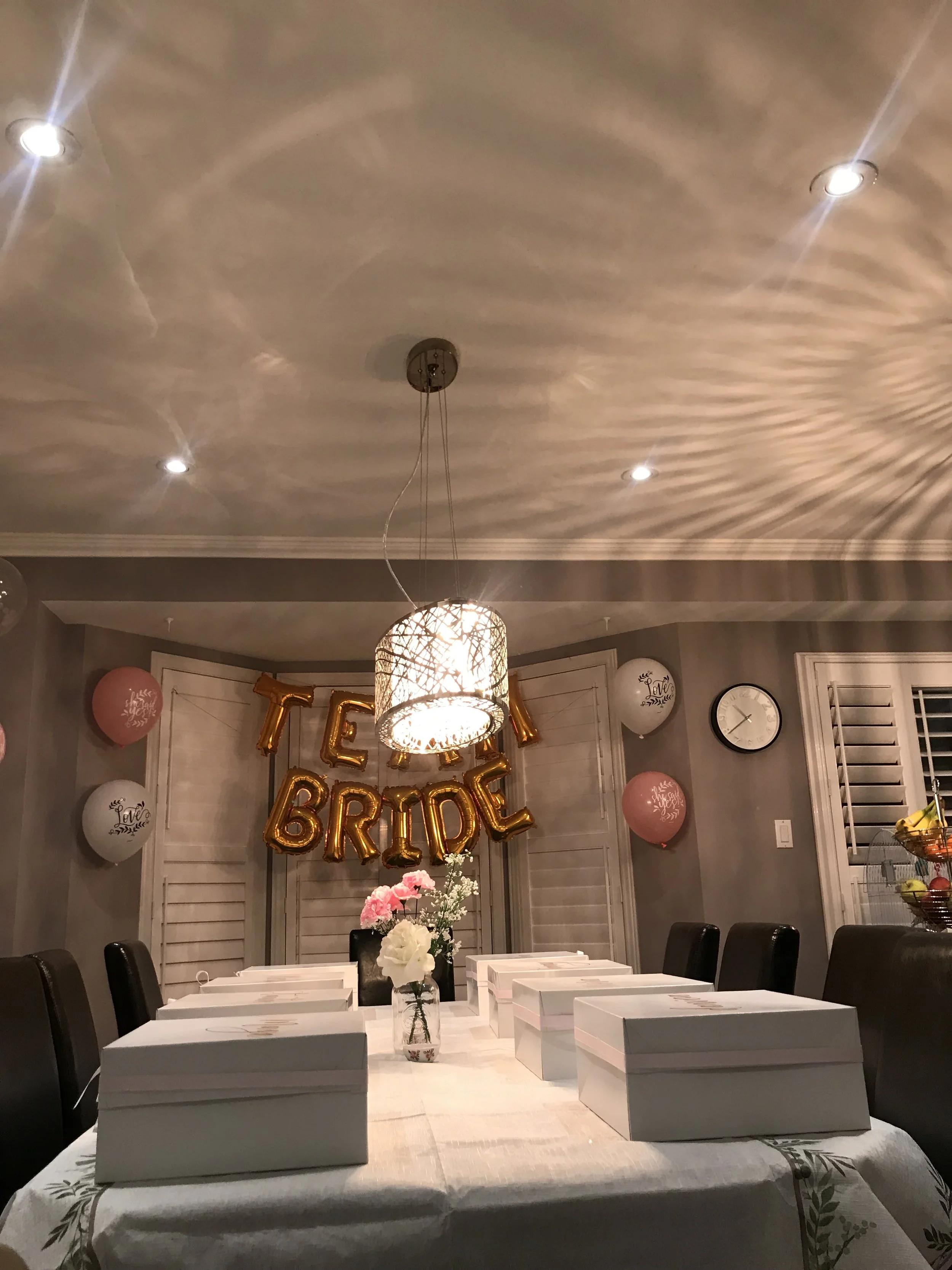 A decorated indoor space with a dining table set for a celebration, featuring white gift boxes, pink and white flowers, and pink balloons. Gold foil balloons spell out 'TEAM BRIDE' on the wall, and there are balloons with the word 'Love' and decorati