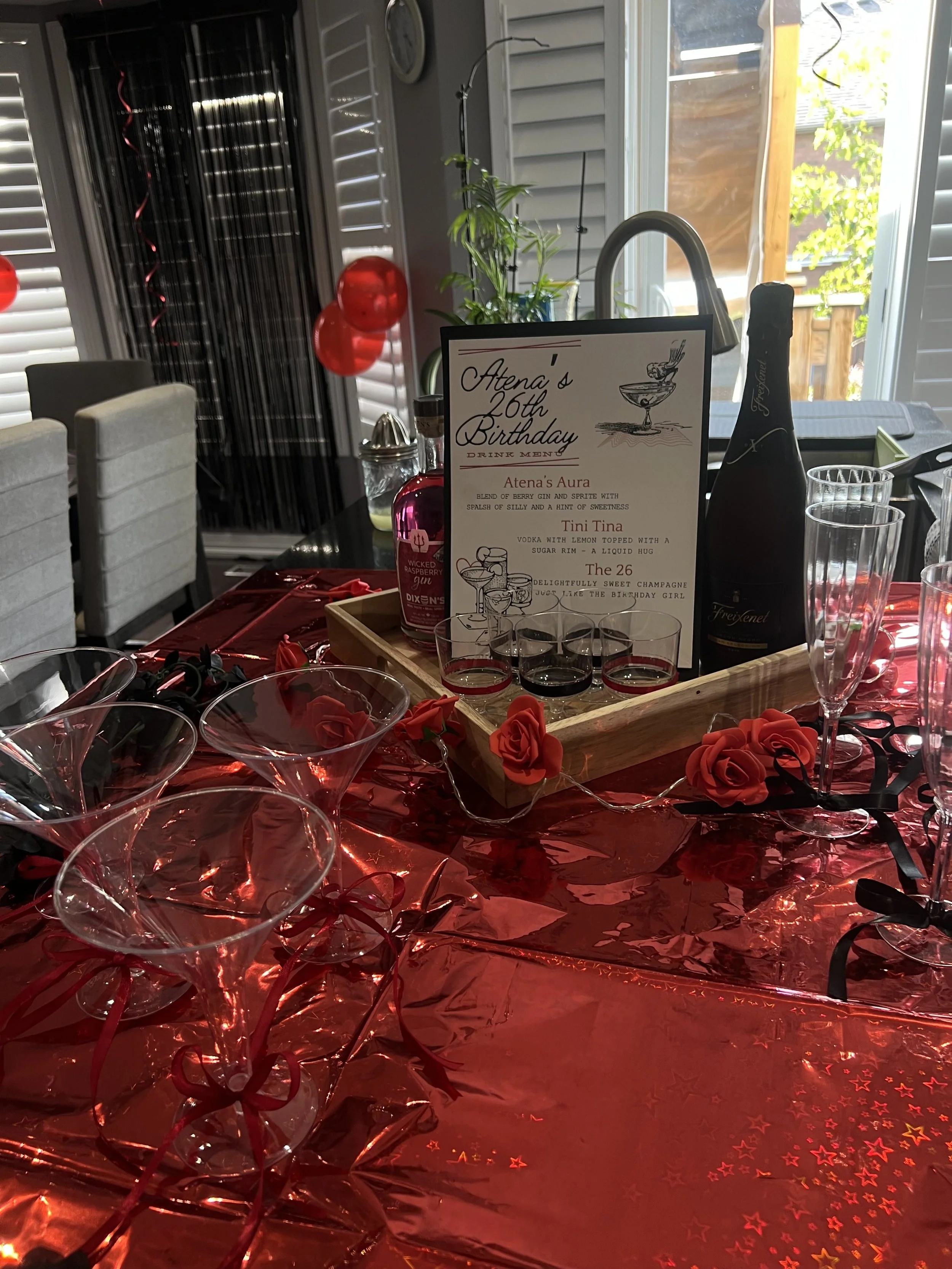 A birthday celebration setup with a red tablecloth decorated with fake roses, string lights, and star-shaped confetti. There is a menu card with birthday drinks, along with glasses, a bottle of champagne, and a bottle of raspberry gin. A sunroom with