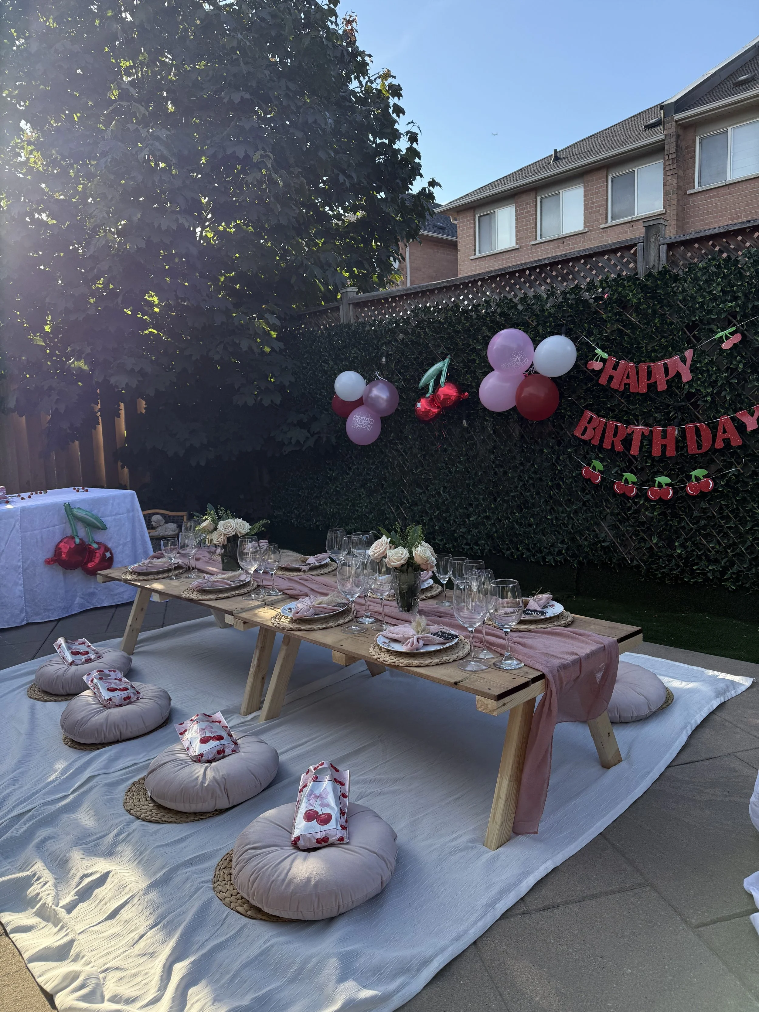 Outdoor birthday celebration setup with a low wooden table decorated with pink table runner, floral centerpiece, and pink napkins; surrounded by beige floor cushions with cherry-patterned cloths, on white blanket, with balloons, "HAPPY BIRTHDAY" bann