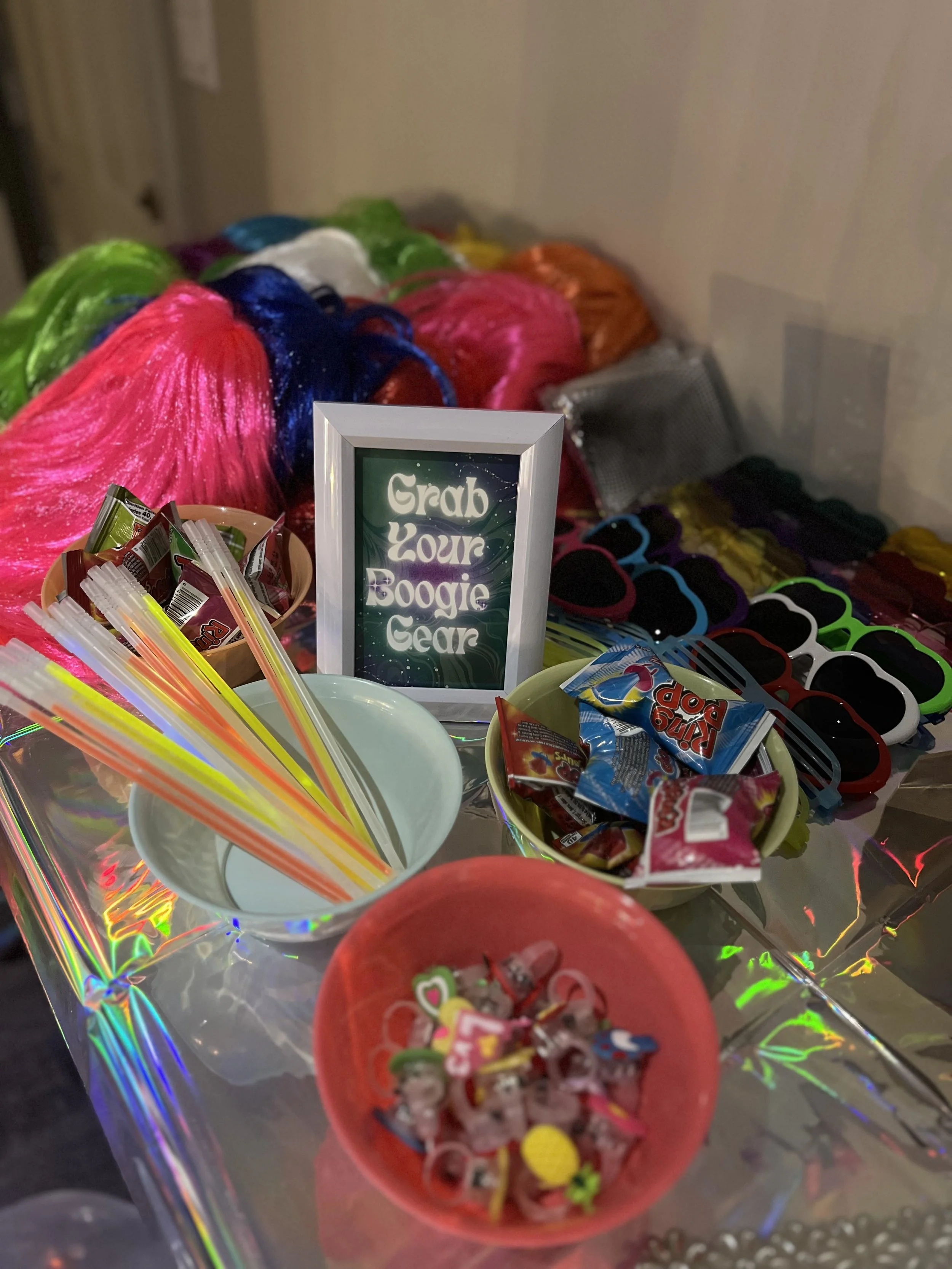 Colorful rainbow of cotton candy, various candies/favors, sunglasses, and a sign that reads 'Grab Your Boogie Gear' at a party or event.