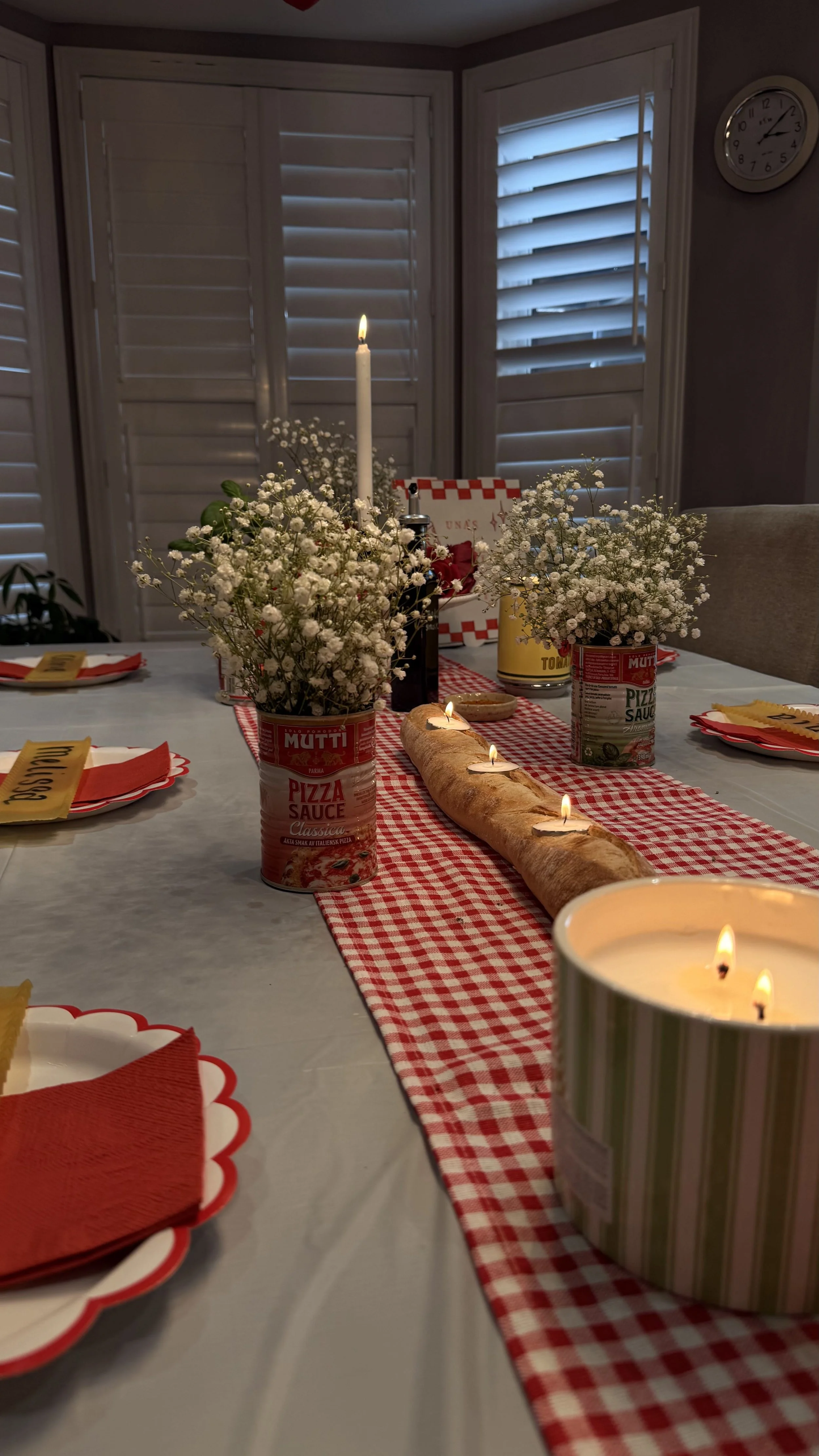 A dining table decorated with red and white checkered table runner, featuring a baguette with candles, flower arrangements in tomato cans, and lit candles, set for a meal.