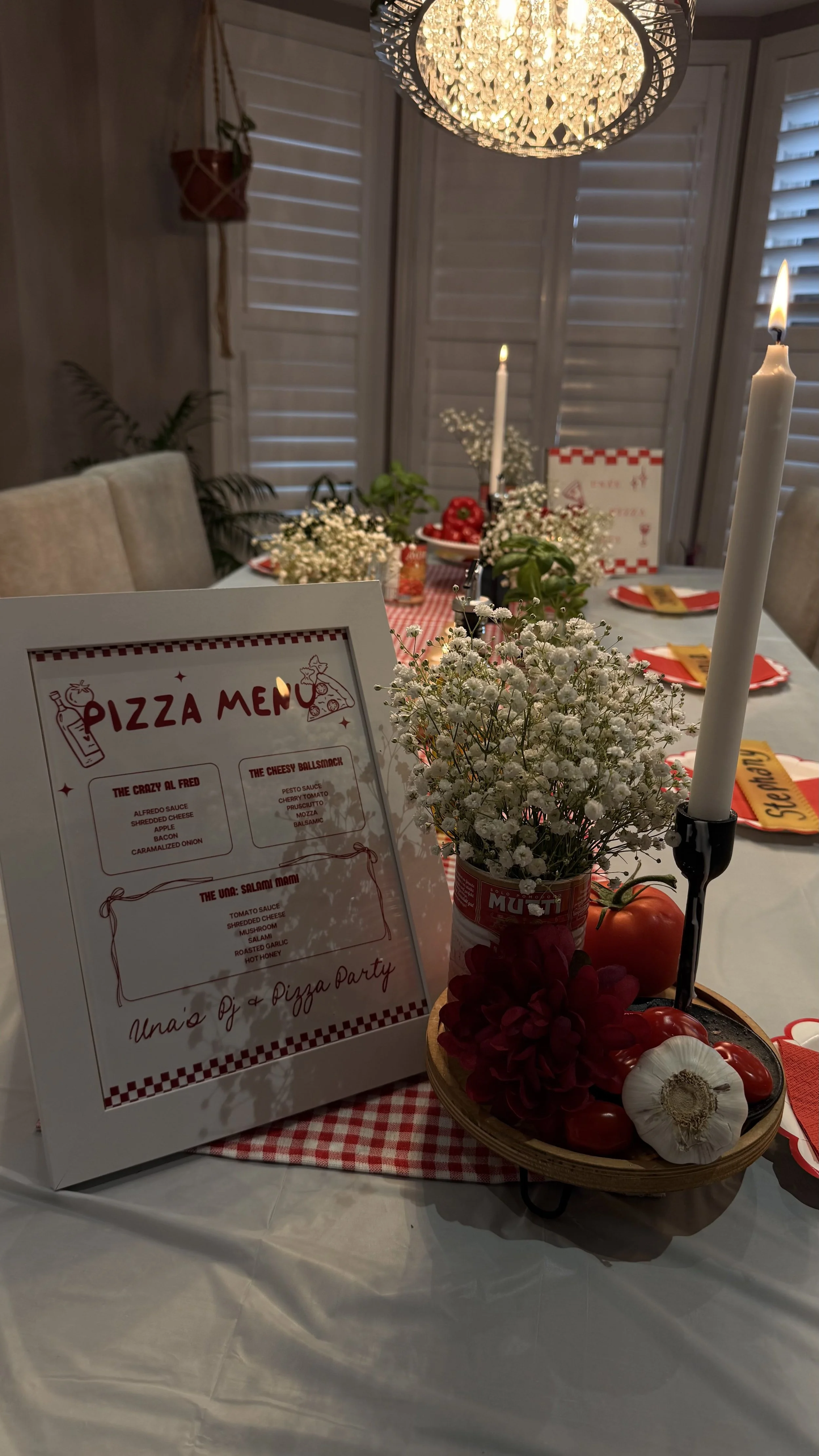 A dining table decorated for a pizza party with a pizza menu, fresh flowers, tomatoes, and candles. The table has a red and white checkered tablecloth, and there's a chandelier hanging above. The background shows window shutters.