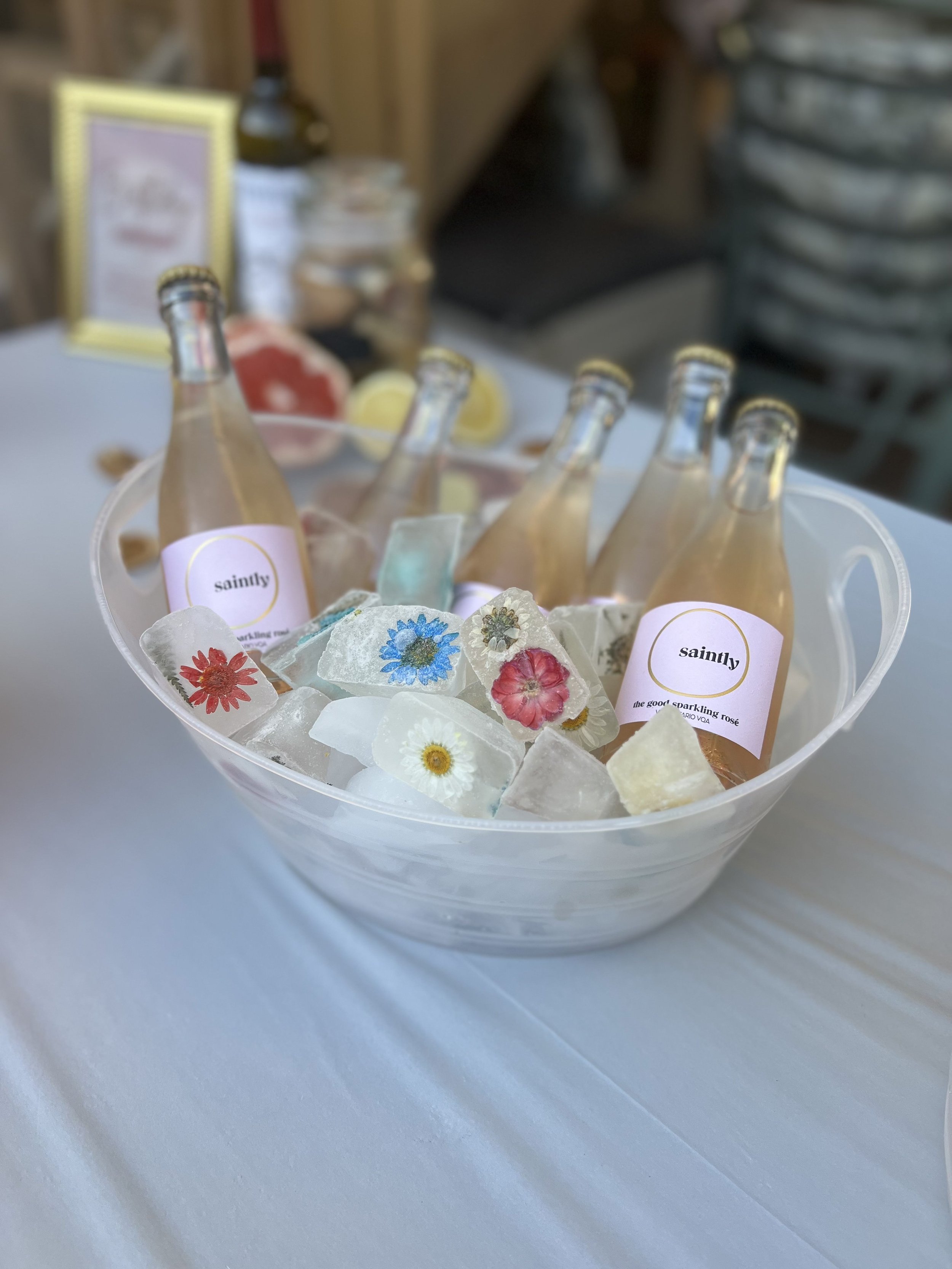 A clear plastic bowl filled with ice cubes and colorful rocks with pressed flowers inside. Several glass bottles with pink and white labels are in the bowl, some filled with a sparkling pink beverage. The background shows a blurry table with some objects and a wooden wall.