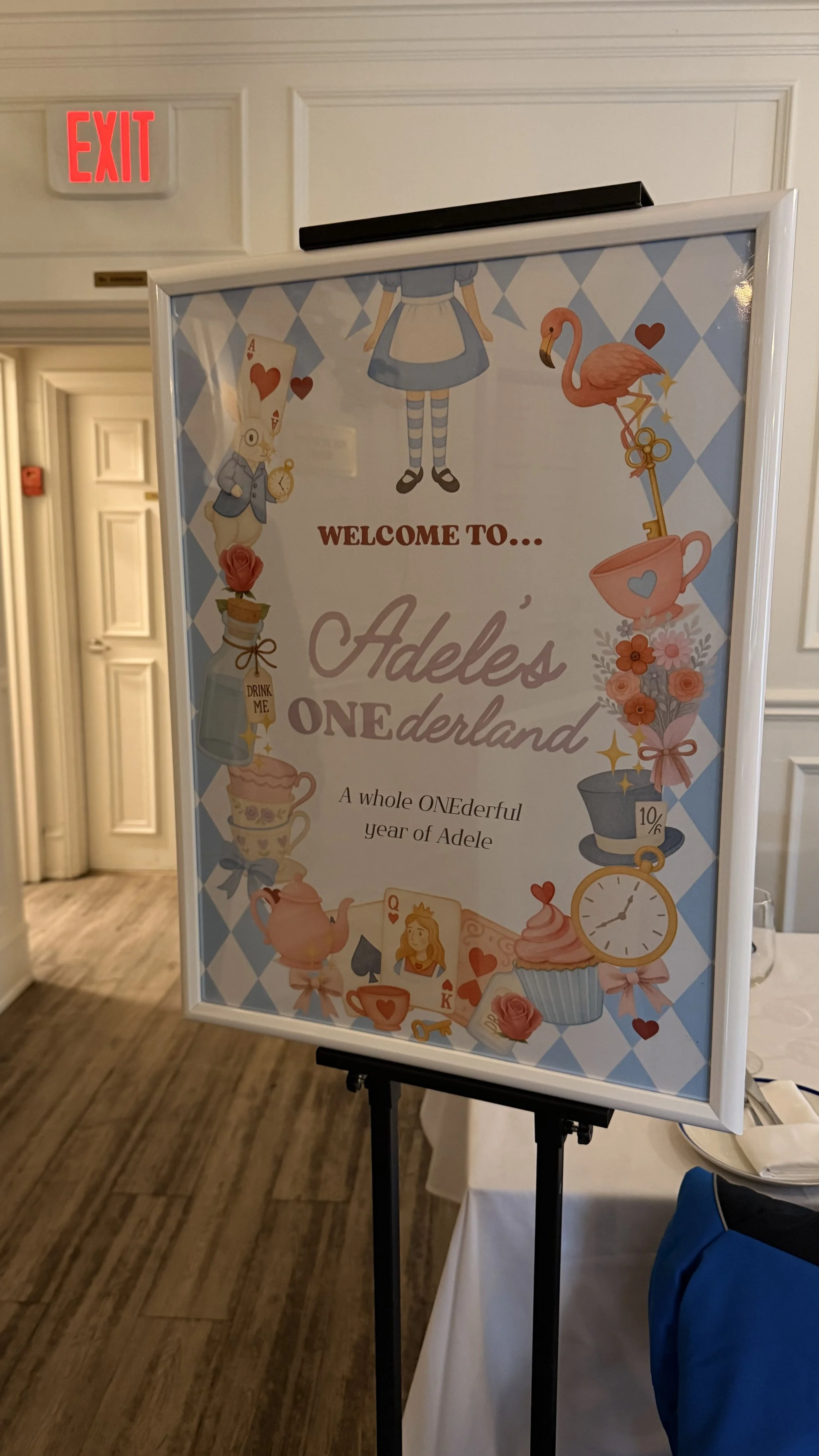 Event sign on a white stand with a decorative Alice in Wonderland theme, including images of characters, teacups, a pocket watch, roses, and playing cards, welcoming guests to Adele's celebration.