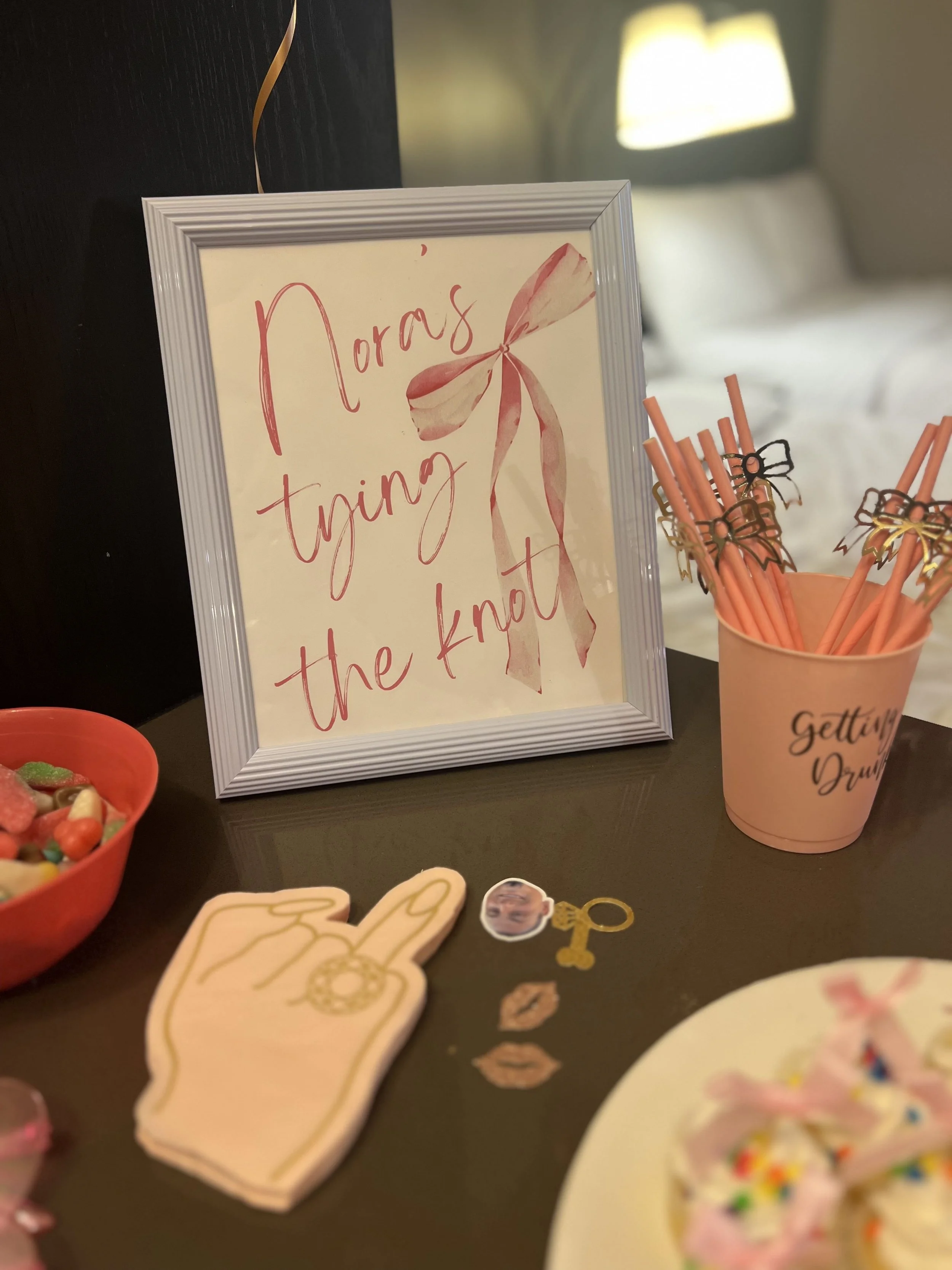 Decorative sign with pink ribbon and handwritten text, surrounded by pink straws in a cup and cookies or decorations on a table in a room with a bed and lamp in the background.