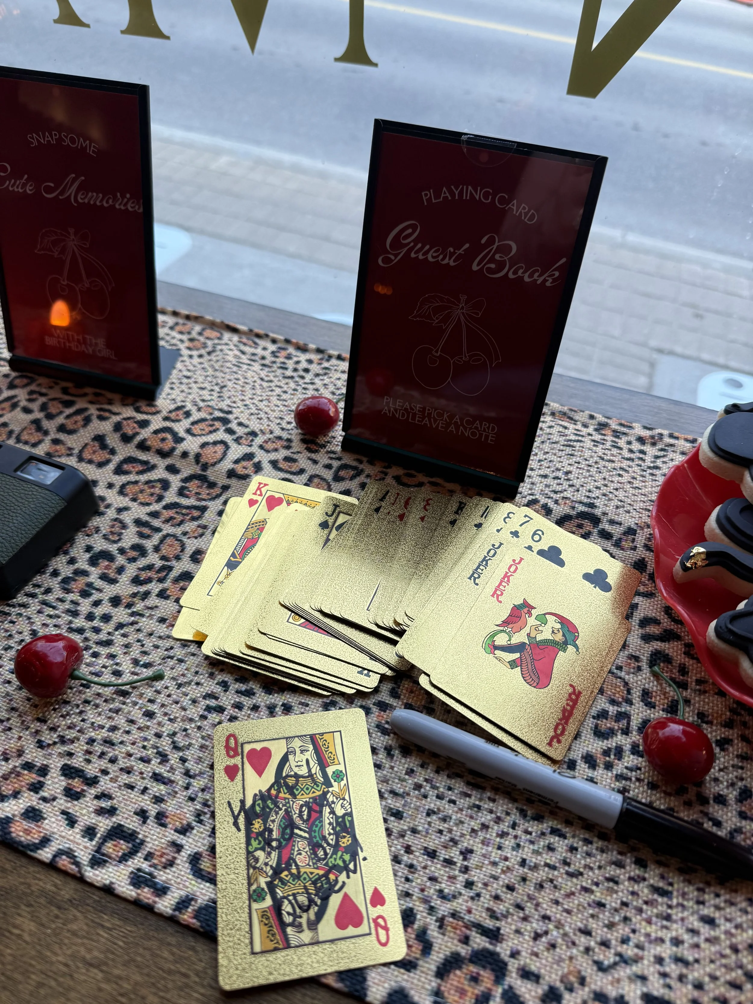 Playing cards, including a Queen of Hearts and a King of Clubs, spread out on a textured fabric tablecloth with cherries and a white marker nearby, set on a table in front of a window.