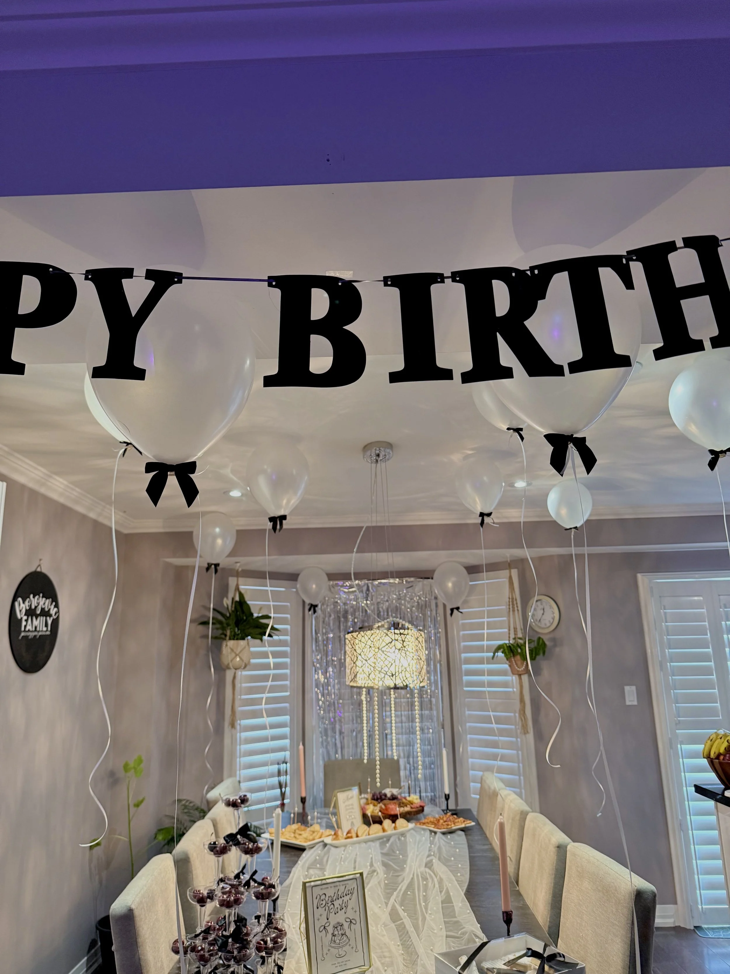 Decorated birthday party with white balloons, black 'Happy Birthday' banner, and a table with various desserts and snacks.
