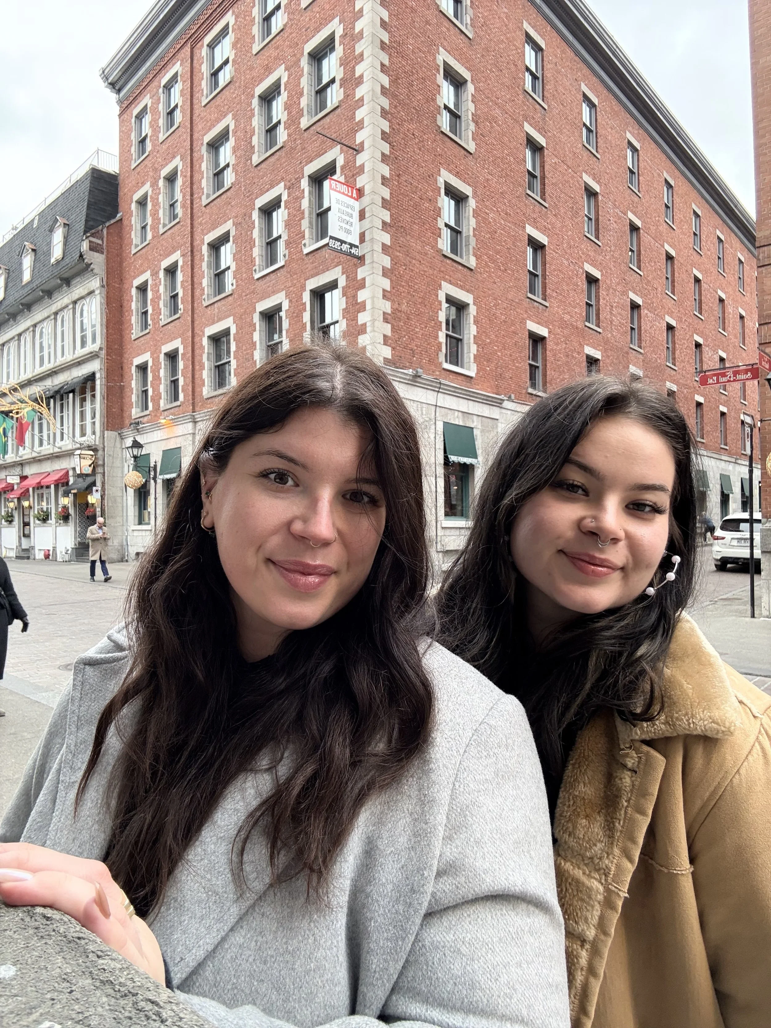 Two young women with dark hair and piercings smiling in an urban setting, with brick buildings and street signs in the background.