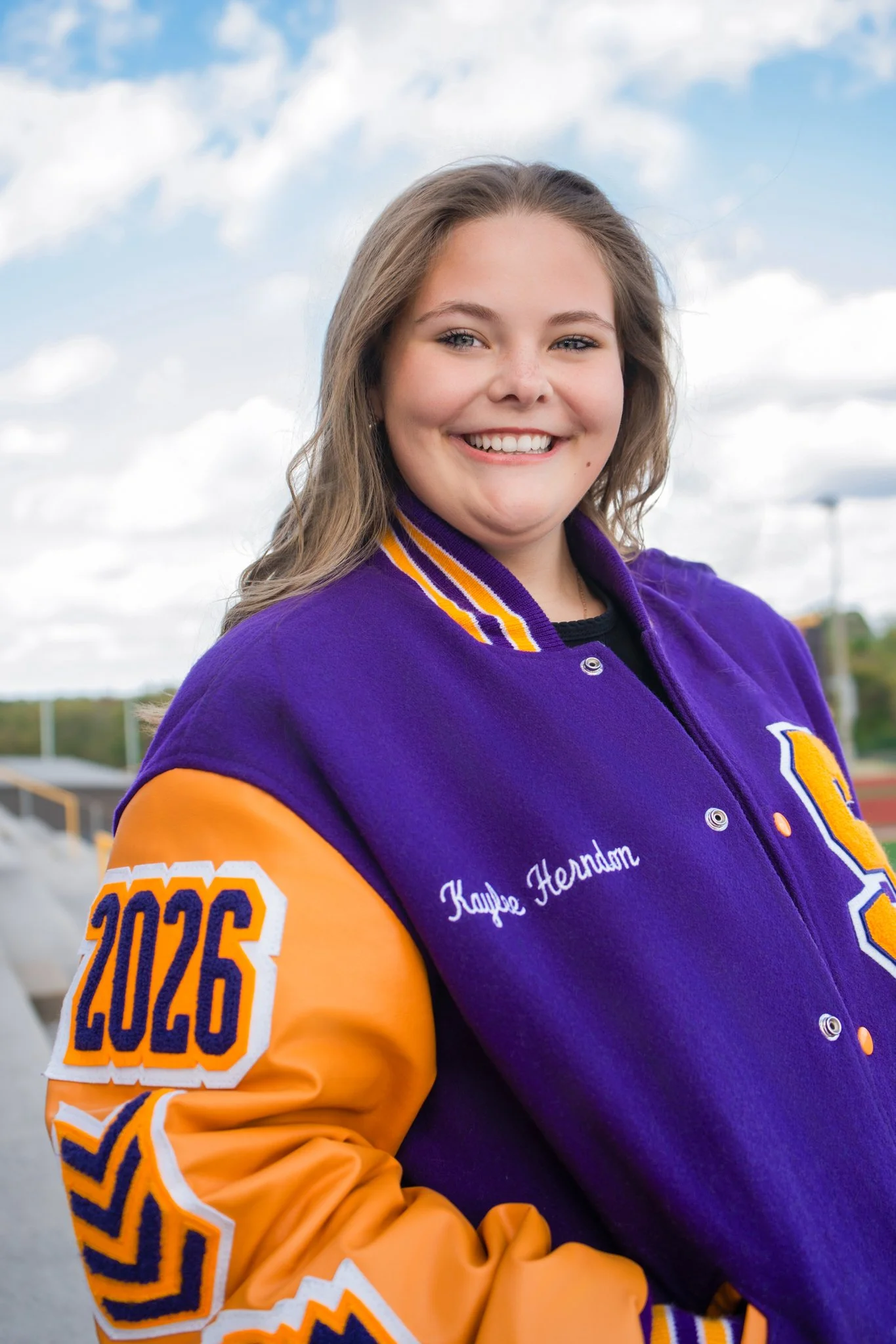 Close-up of a young woman with long light brown hair, smiling, wearing a purple and orange letterman jacket with the year 2025 on the sleeve and a name embroidered on the chest, standing outdoors with a cloudy sky in the background.