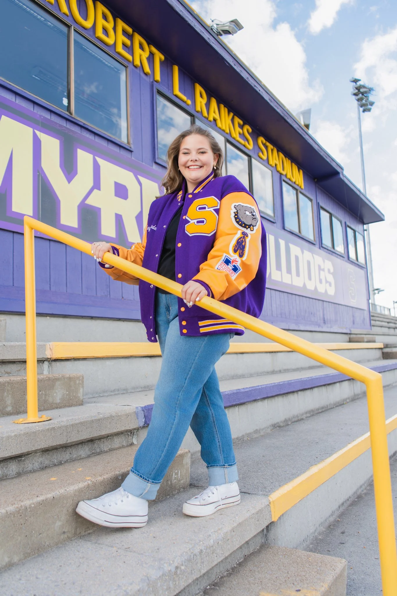 A young woman with wavy brown hair smiling at the camera, wearing a purple and yellow varsity jacket with a bulldog patch and a large letter 'S', light blue jeans with rolled cuffs and white platform sneakers, standing on concrete steps outside a pur