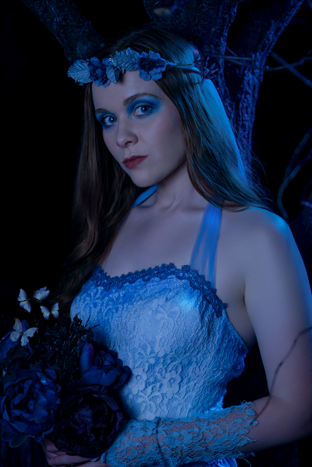 A woman with blue eyes and long brown hair wearing a white lace dress and a floral crown, standing in a dark outdoor setting with blue lighting and holding a bouquet of dark roses and butterflies.