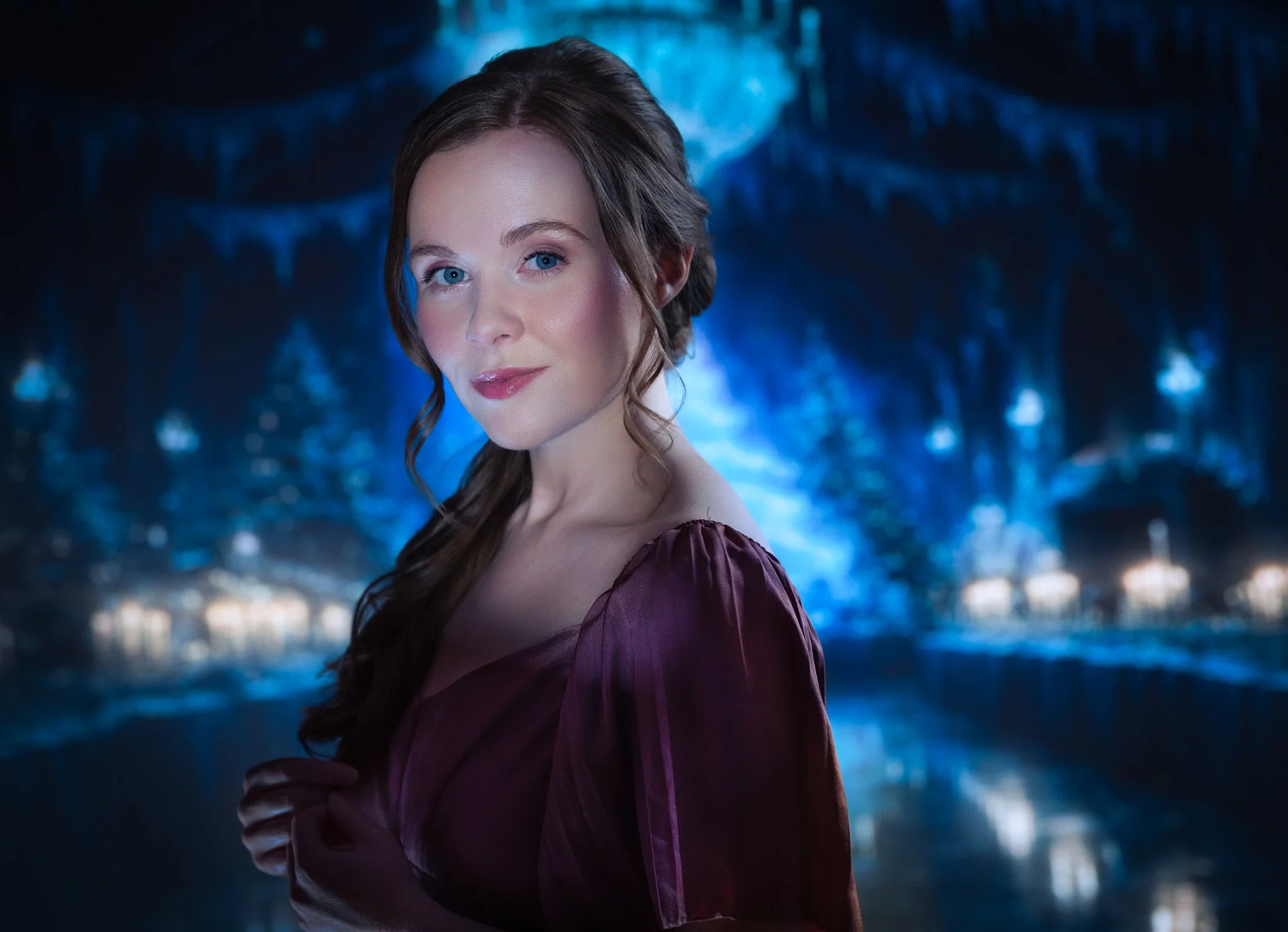A woman with long brown hair and blue eyes, wearing a purple dress, standing in front of icy blue cave walls.