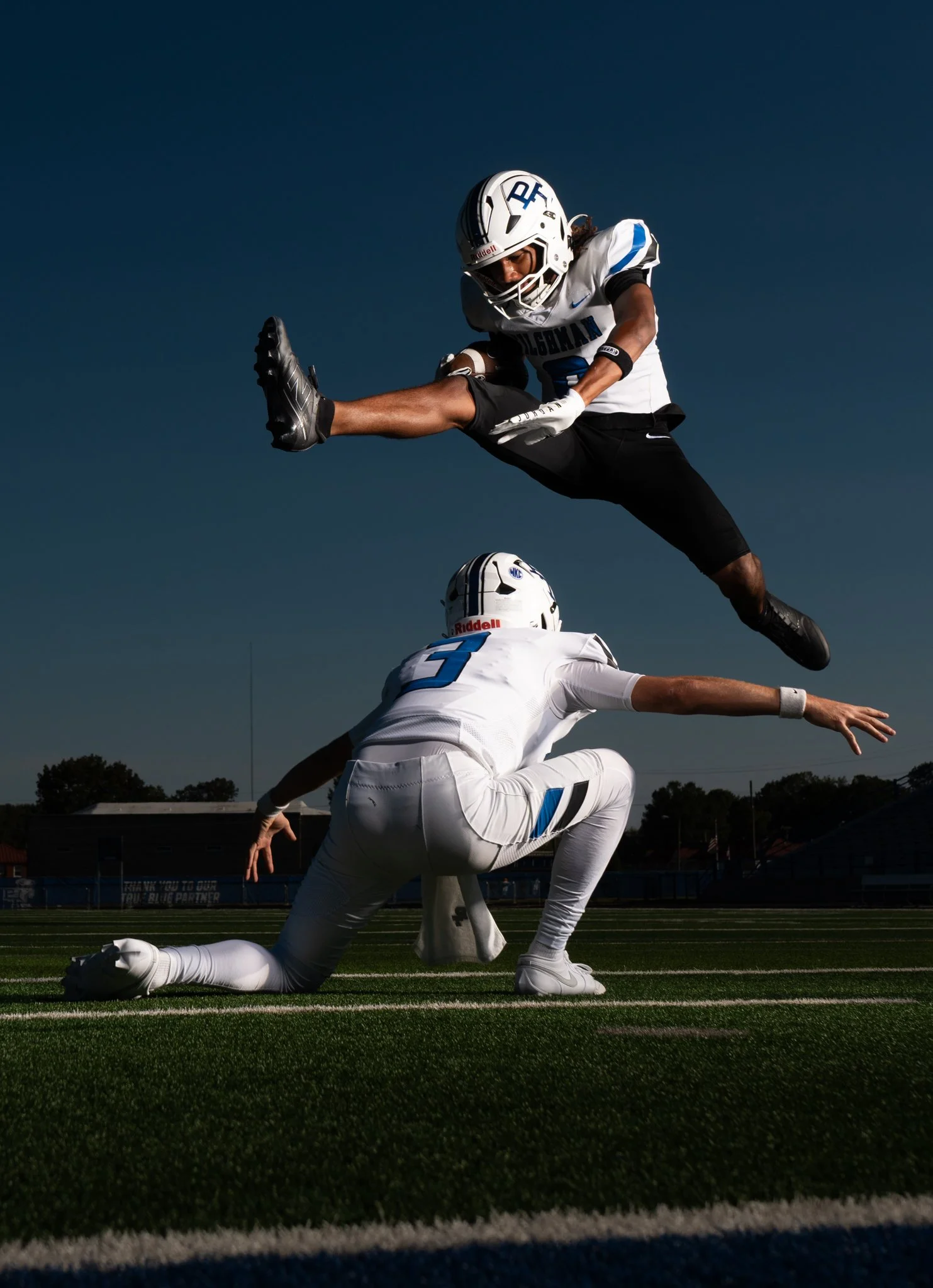 A football player in white uniform is diving to catch a football in midair, while another player in white is crouching on the field, both under a dark blue sky.