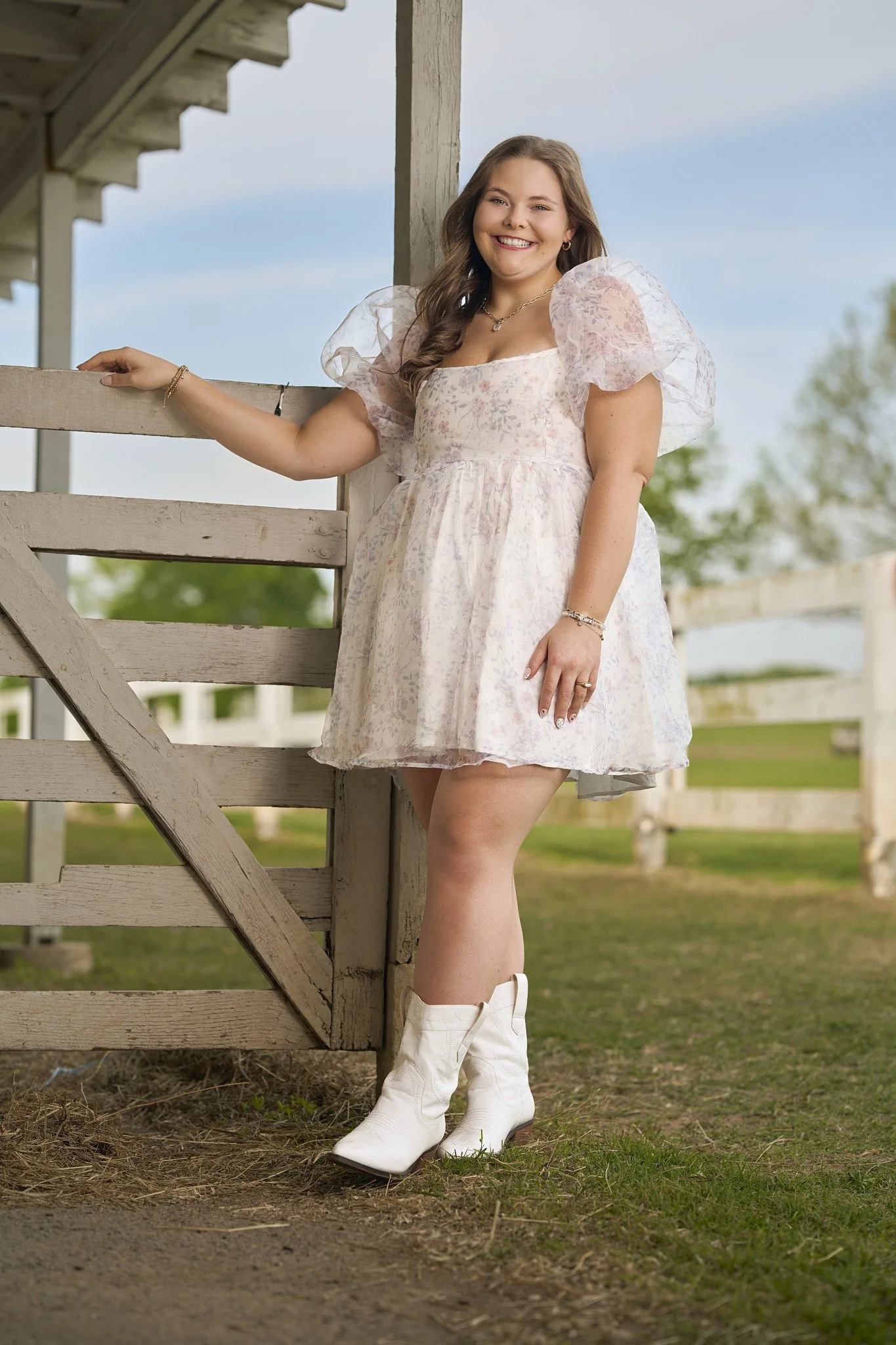 A young woman wearing a pink floral dress and white cowboy boots standing outdoors near a wooden fence, smiling and posing for the photo.