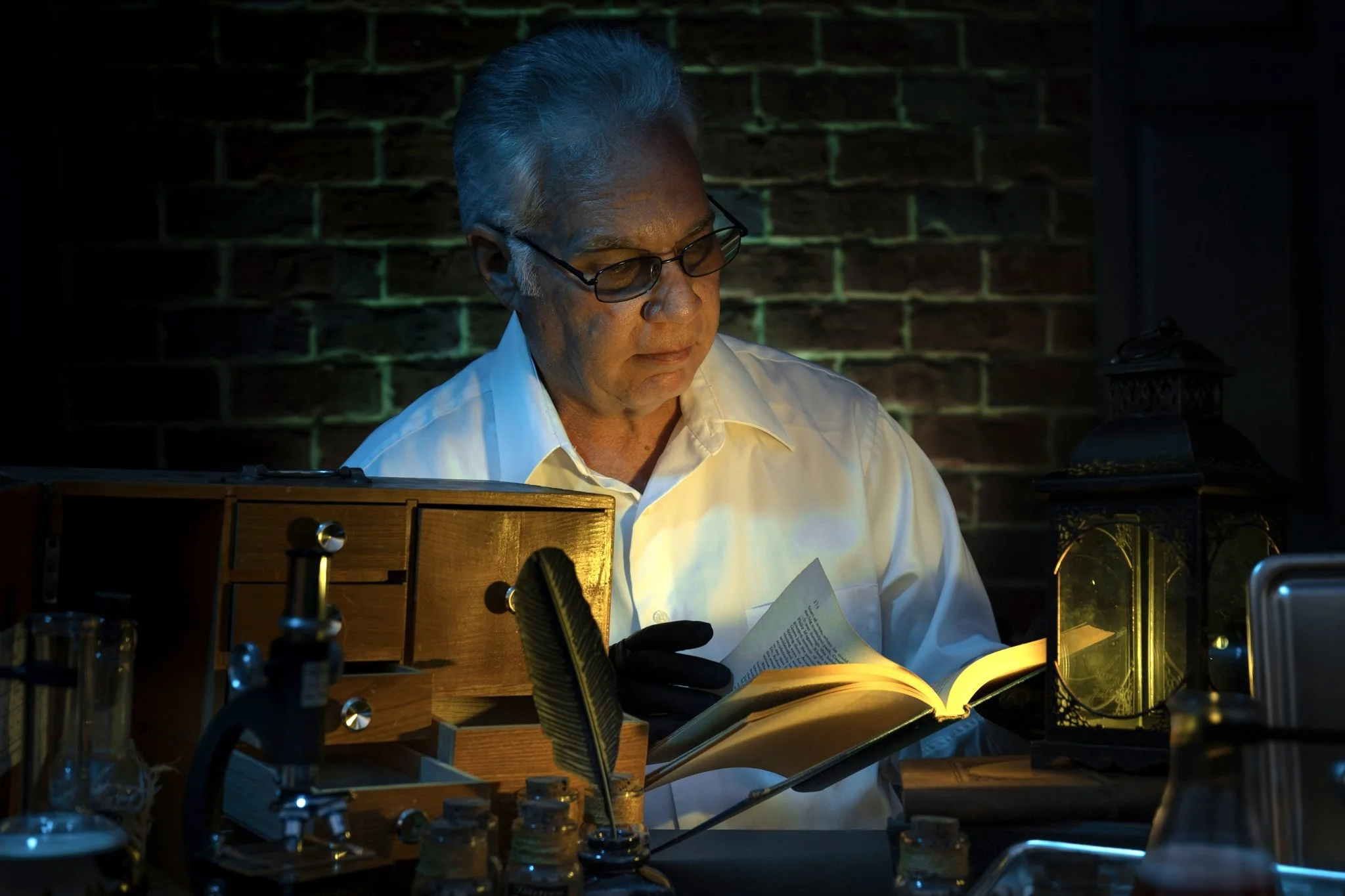 A man wearing glasses and a white shirt is working in a dimly lit laboratory with various scientific equipment and a brick wall in the background. He is reading a document illuminated by a desk light.