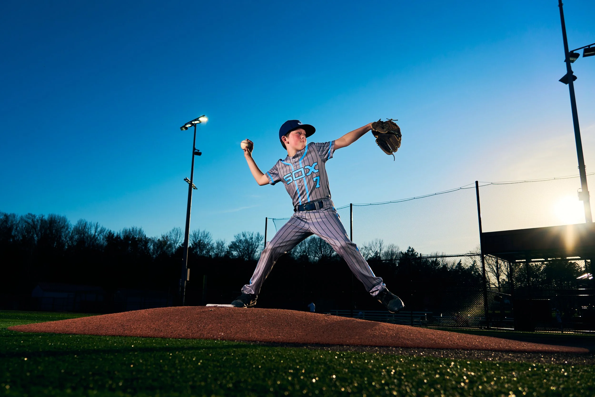 A young boy in a baseball uniform pitching on a baseball field during sunset, with bright blue sky and stadium lights in the background.