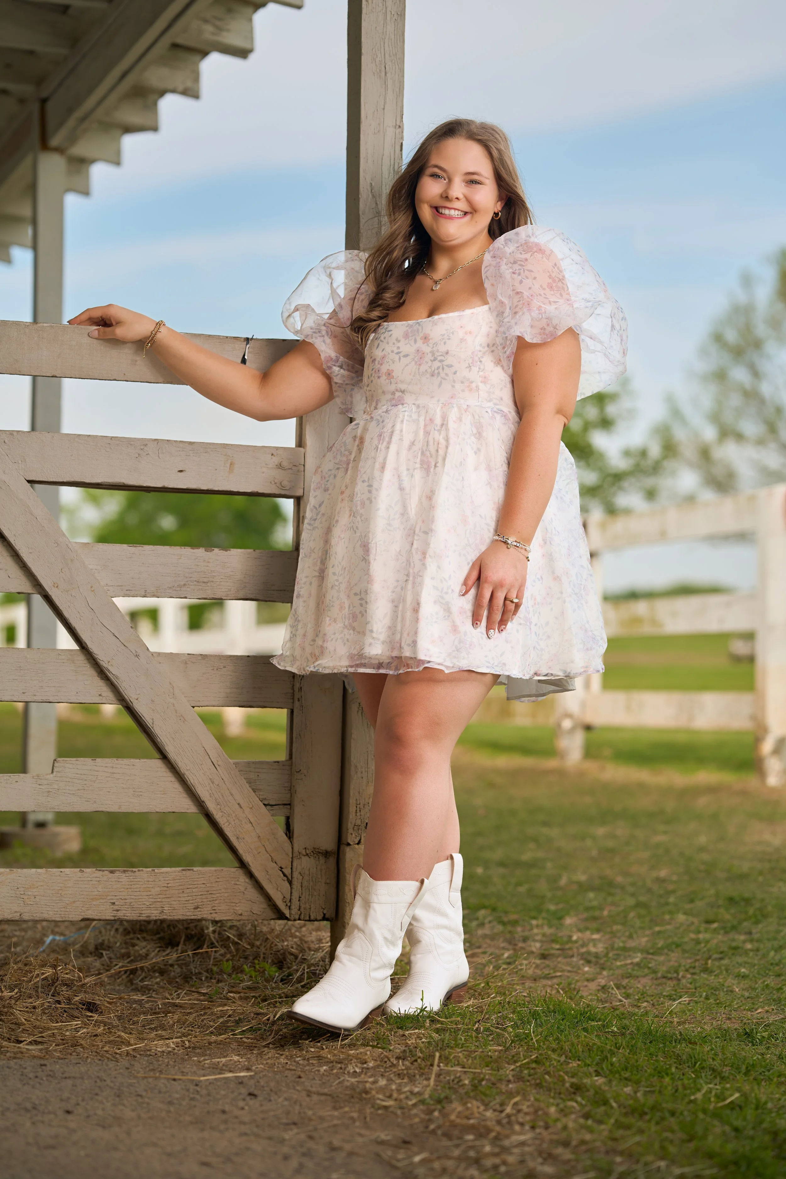 A young woman with long hair smiling and wearing a light-colored floral dress, white cowboy boots, standing outdoors near a wooden fence with clear blue sky and green trees in the background.