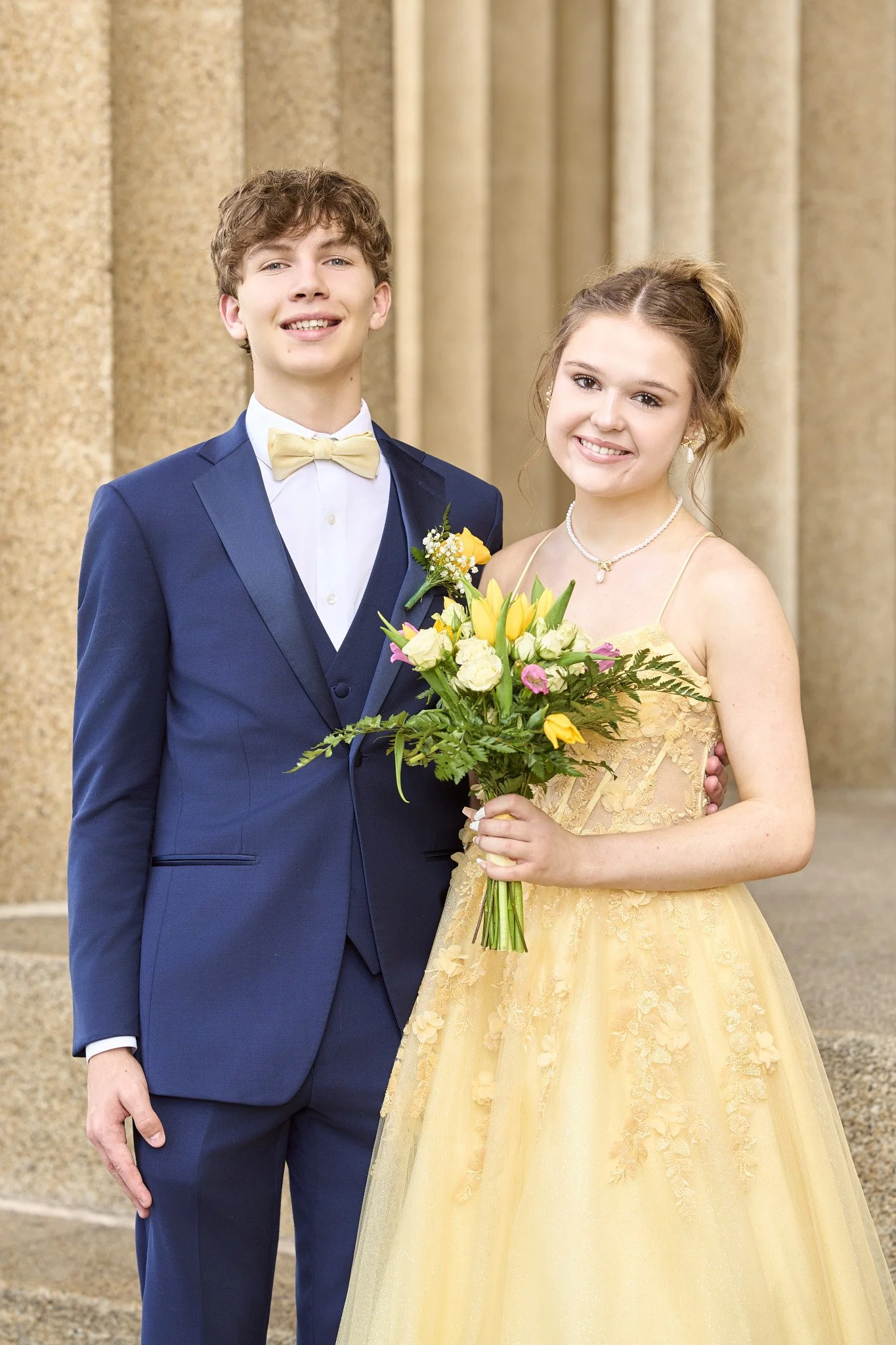 A young man and woman dressed in formal attire, standing outdoors in front of a stone building. The man is wearing a navy blue suit with a beige bow tie, and the woman is in a yellow lace gown holding a bouquet of flowers.