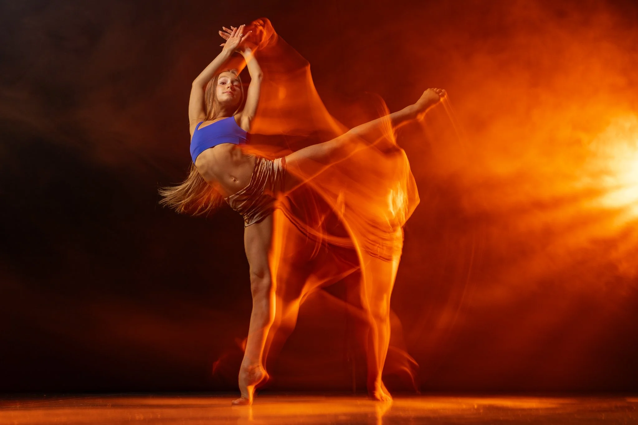A young female dancer in a blue top and striped shorts performs a dance move on a dark stage with orange flames background effects.