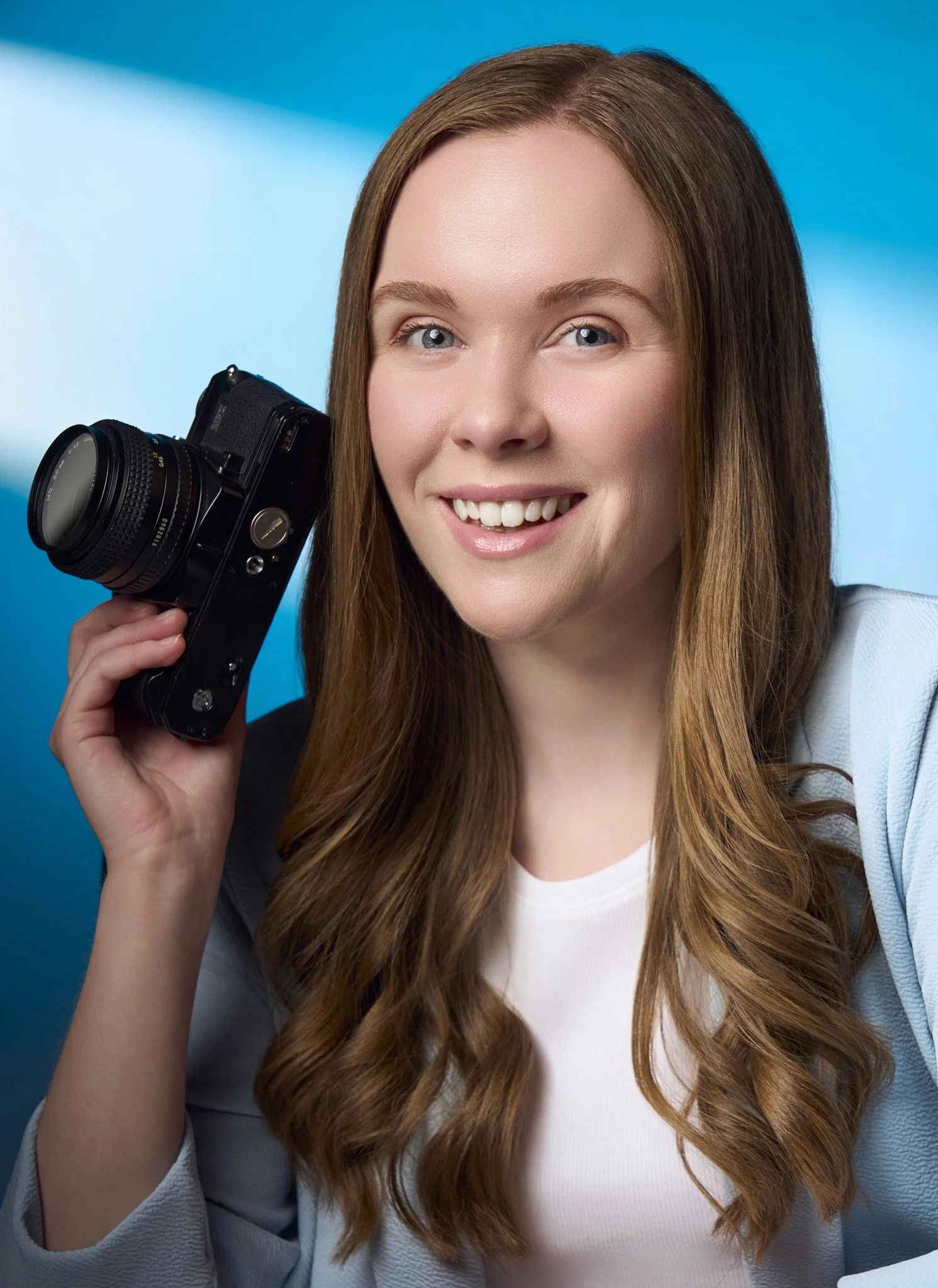 A young woman with long, wavy auburn hair holding a vintage black camera to her ear, smiling against a blue background.