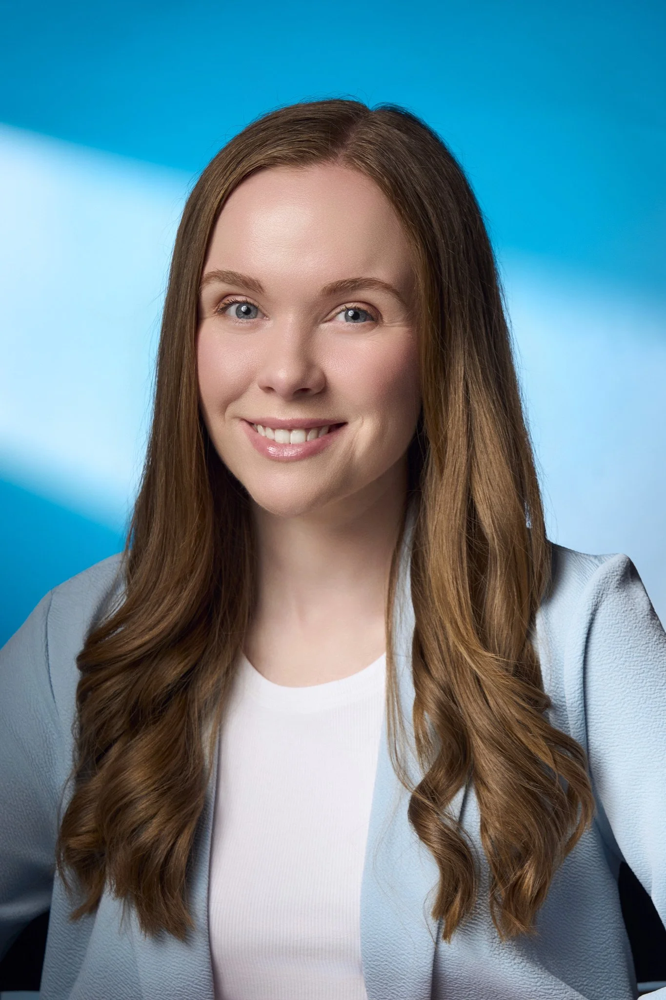 Close-up portrait of a young woman with long, wavy brown hair, blue eyes, and fair skin, smiling softly, wearing a light-colored blazer and a white top, against a bright blue background.