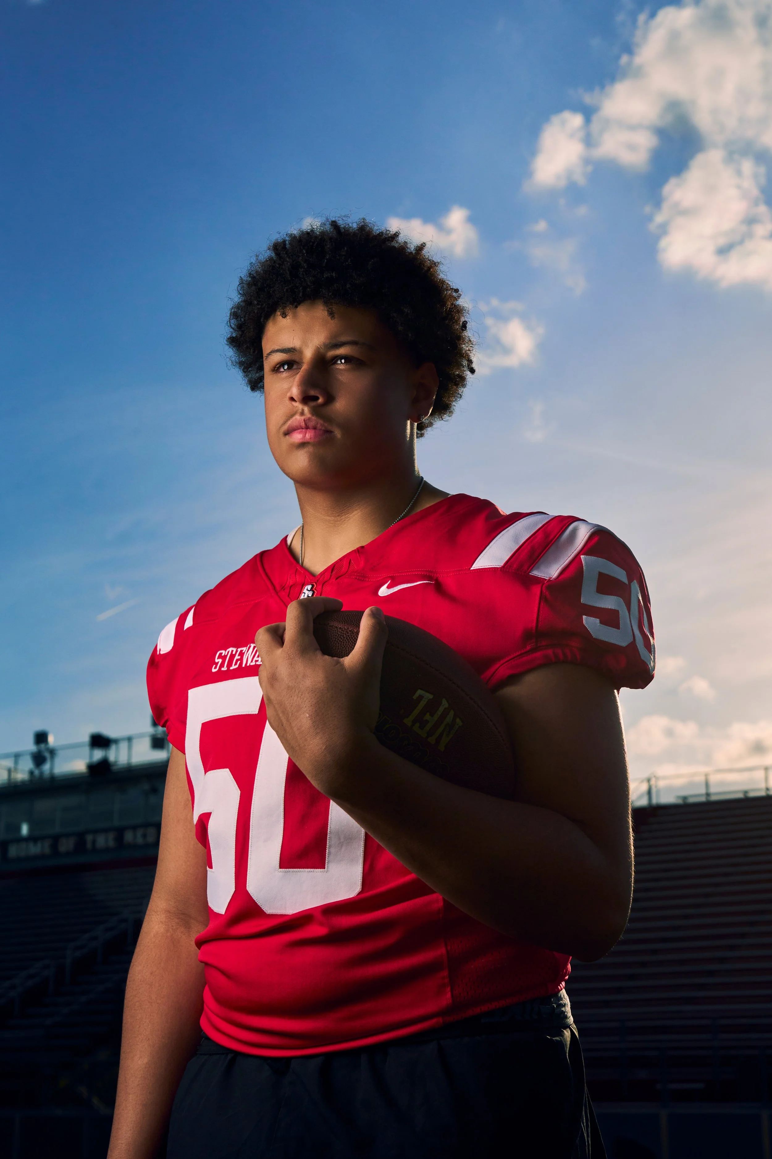 A young male football player wearing a red jersey with the number 88, holding a football against a sunset sky with clouds, at a stadium.
