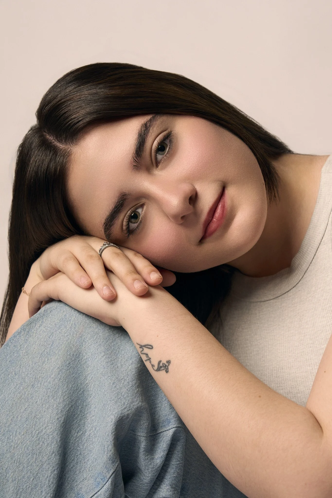 Close-up of a young woman with shoulder-length dark hair, resting her head on her hand, with a gentle smile, wearing a light-colored shirt and a gray cloth, with a tattoo on her forearm.