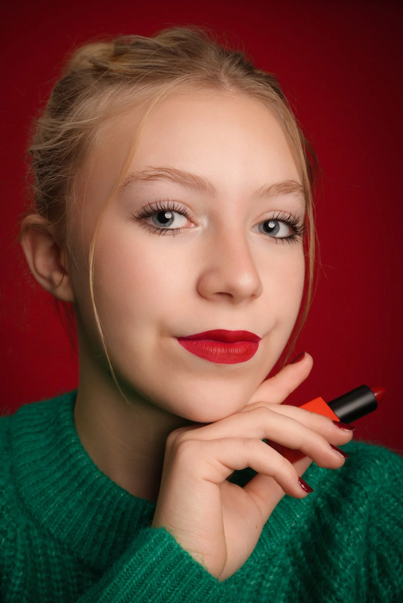 Close-up of a young woman with blonde hair, blue eyes, red lipstick, wearing a green sweater, holding red lipstick, against a red background.