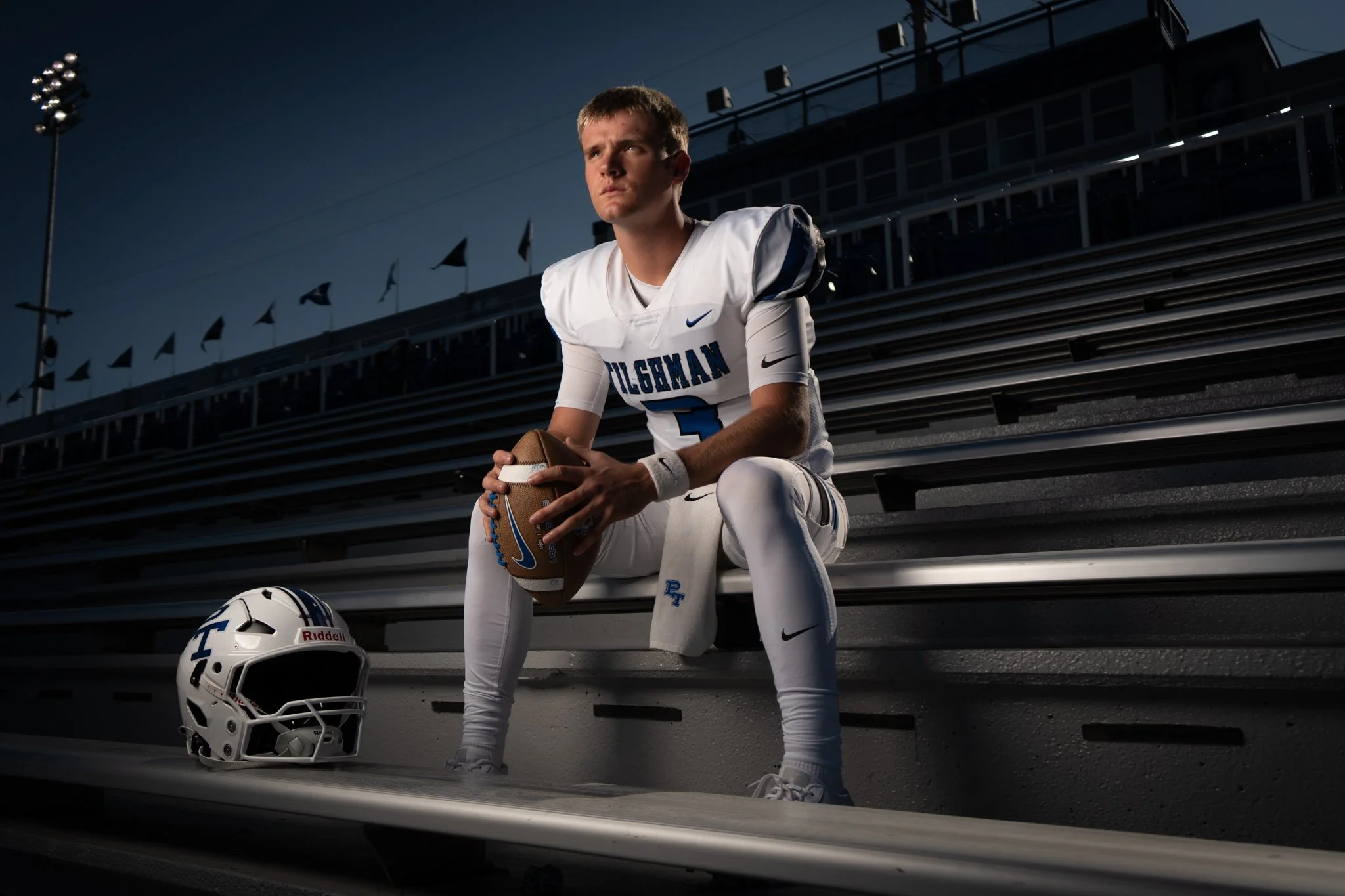 A football player in a white and blue uniform sitting on stadium bleachers, holding a football. A helmet and cleats are placed on the ground nearby. The scene is set at dusk with stadium lights in the background.