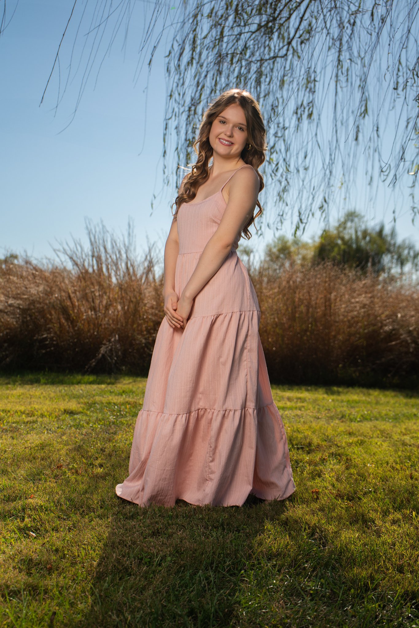 A young woman standing outdoors on grass, wearing a long pink dress with spaghetti straps, with a background of bushes, trees, and a clear blue sky, smiling at the camera.