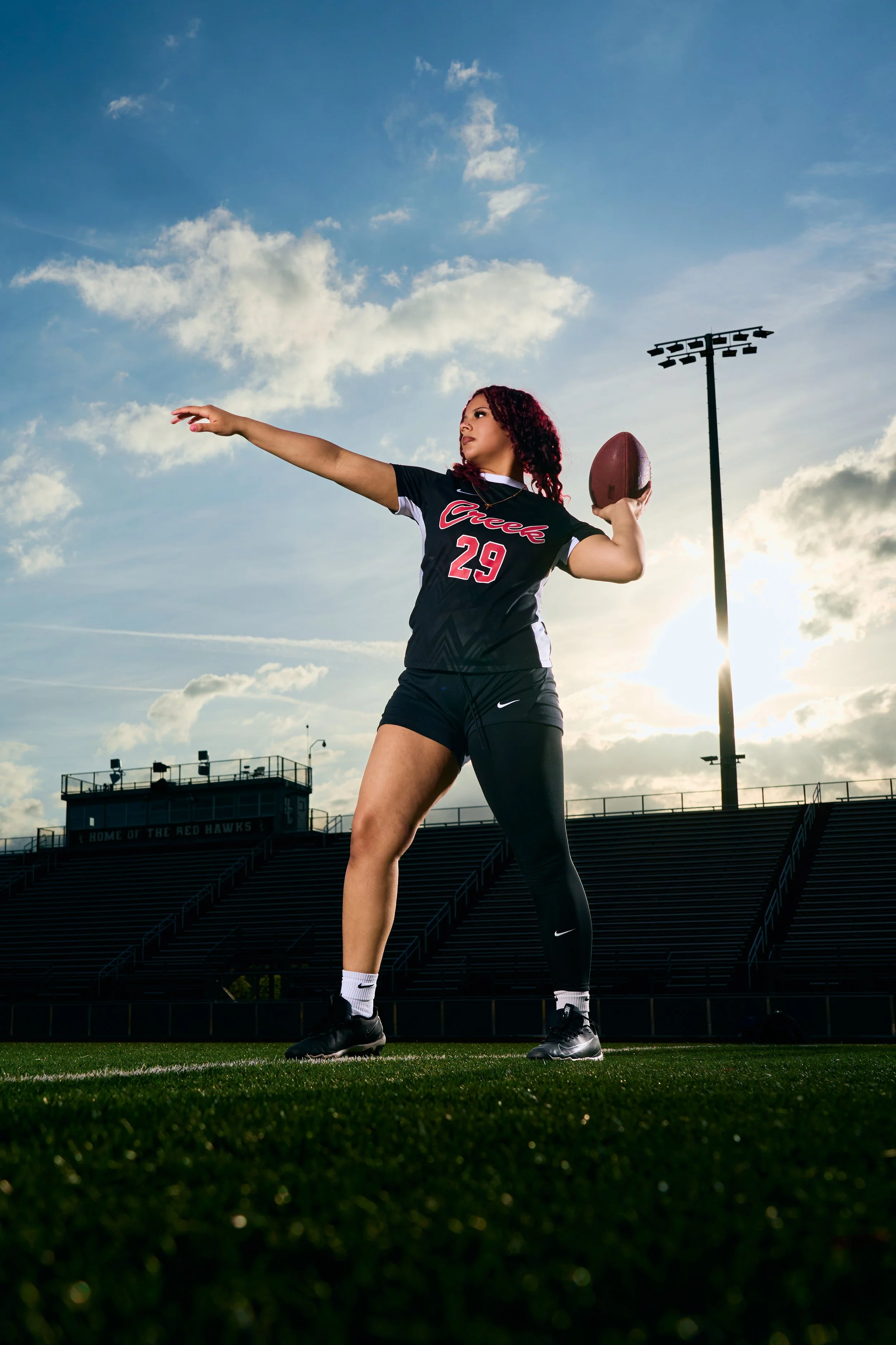 A female football player in a black jersey with the number 29, holding a football in her right hand, standing on a football field with stadium lights and bleachers in the background, during sunset or late afternoon.