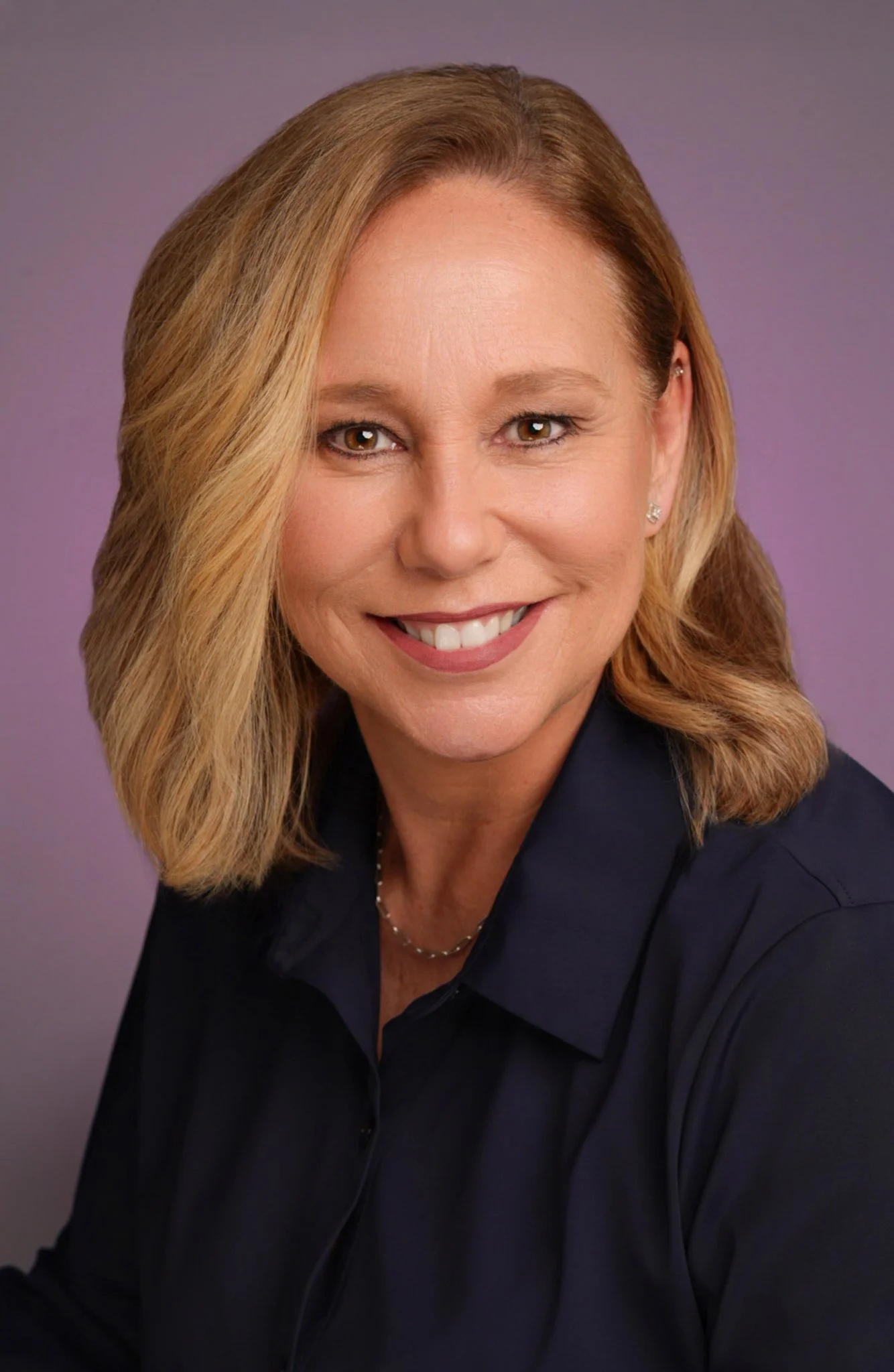 A woman with shoulder-length blonde hair smiling, wearing a dark navy or black collared shirt and a necklace, against a purple background.